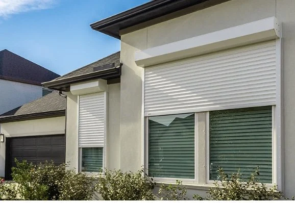 The front of a house with two windows, each with white rolling metal shutters partially closed, and a shingle roof. There are bushes in the foreground.