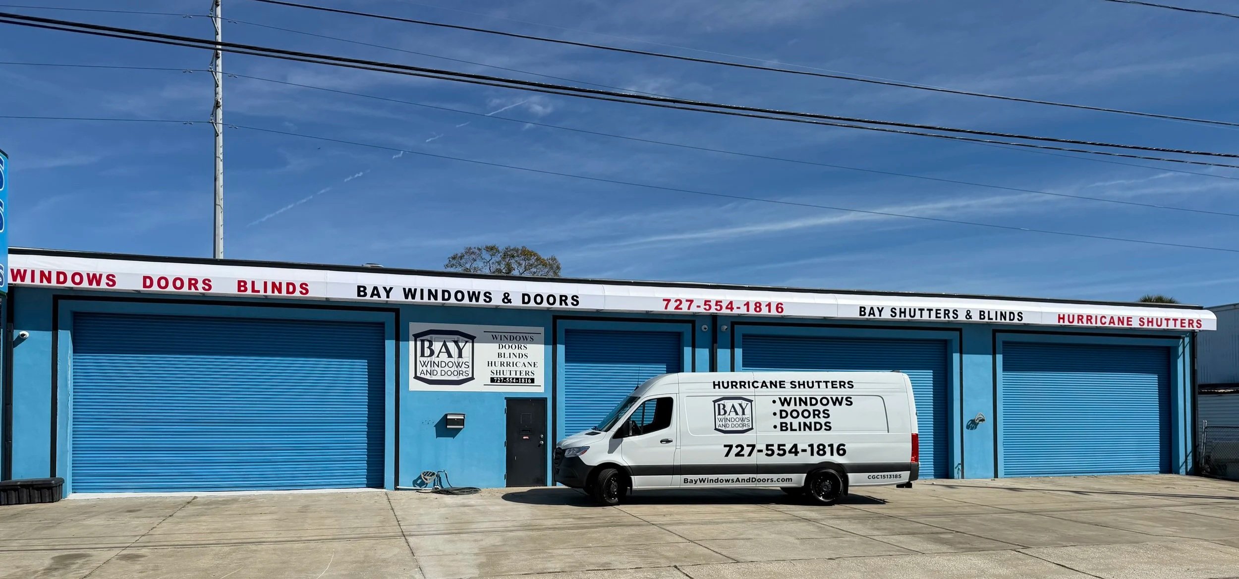 Storefront building with blue roll-up doors, signage indicating it sells windows, doors, blinds, and hurricane shutters. White van parked in front displays the store's services and contact number. Clear sky with power lines above.