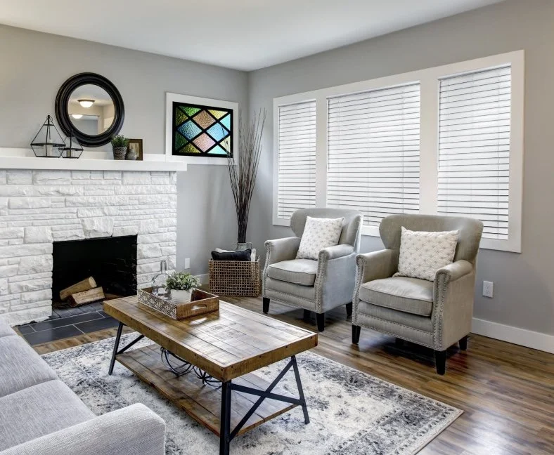 A living room with a white brick fireplace, a wooden coffee table, a gray sofa, two gray armchairs with white pillows, a patterned rug, and three large windows with white blinds.