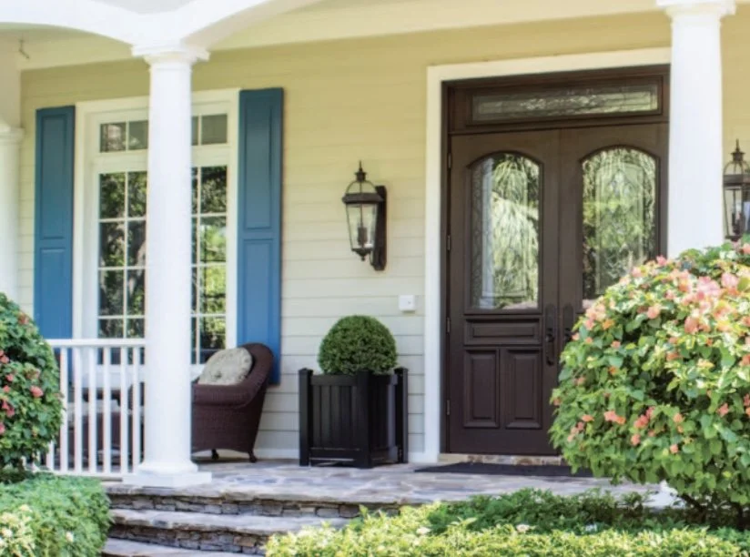 Front porch of a house with a dark brown door, two white columns, blue shutters, a wicker chair, a black planter with a round topiary, and flowering bushes.