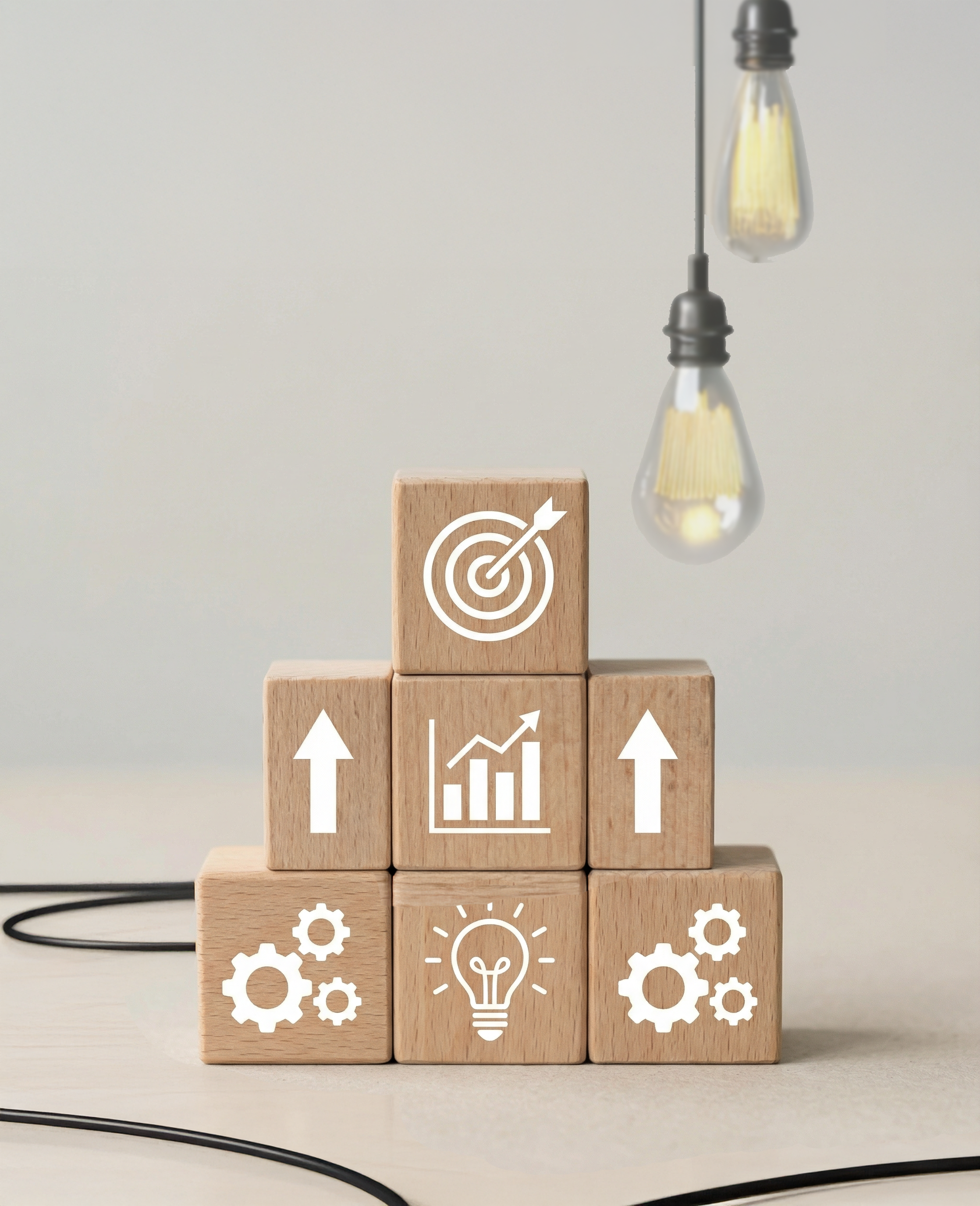 Wooden blocks stacked in a pyramid with icons of gears, a light bulb, upward arrows, a chart, and a target, symbolizing productivity, innovation, and goal achievement, with two hanging light bulbs on the right.