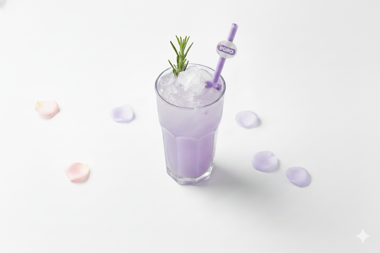 A tall glass of a lavender-colored beverage topped with crushed ice, garnished with a sprig of rosemary, with a purple straw labeled "BUBO". Flower petals are scattered around the glass on a white surface.