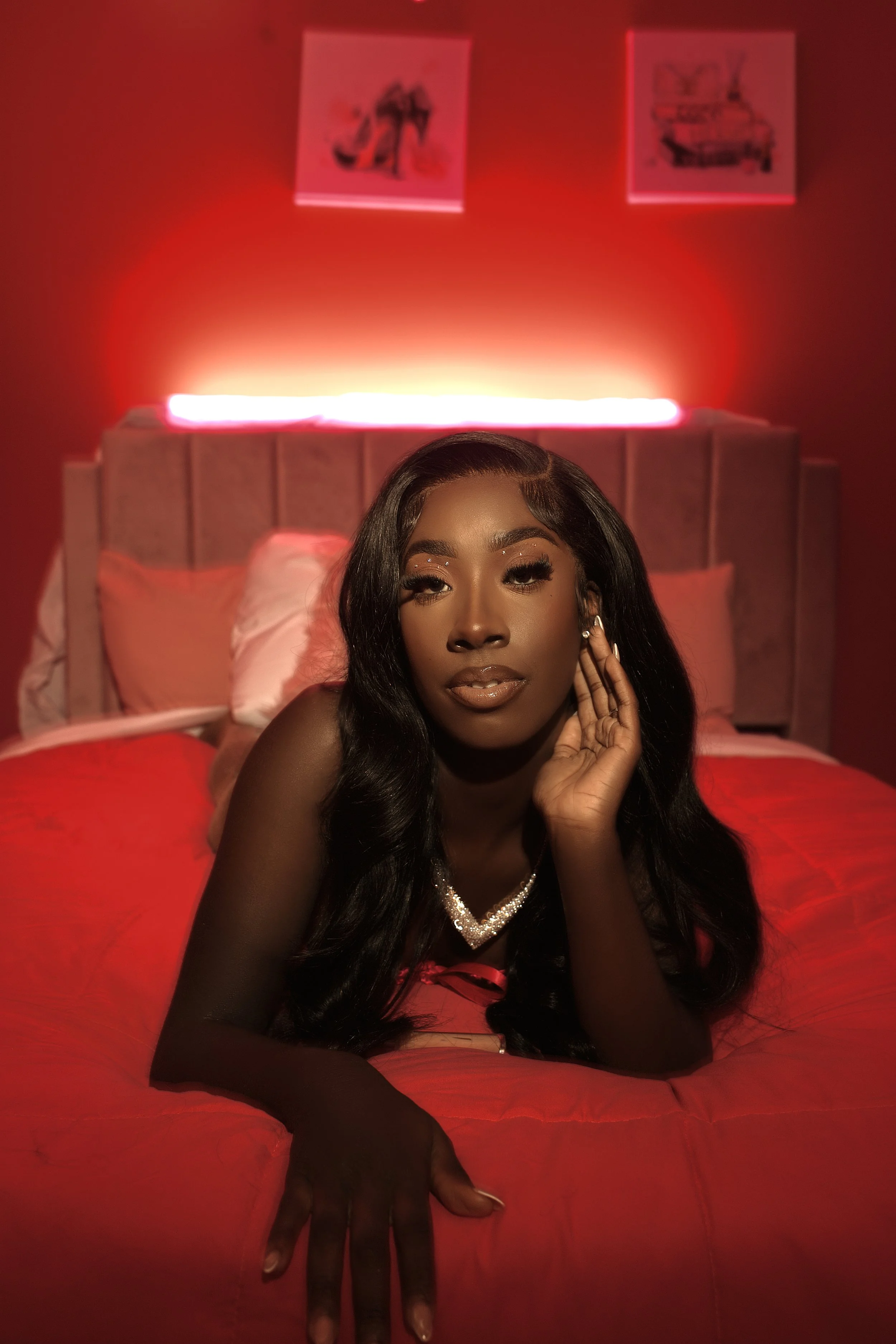 Young woman with long black wavy hair, wearing jewelry, lying on a red bed in a dimly lit room with red lighting and two framed pictures on the wall behind her.