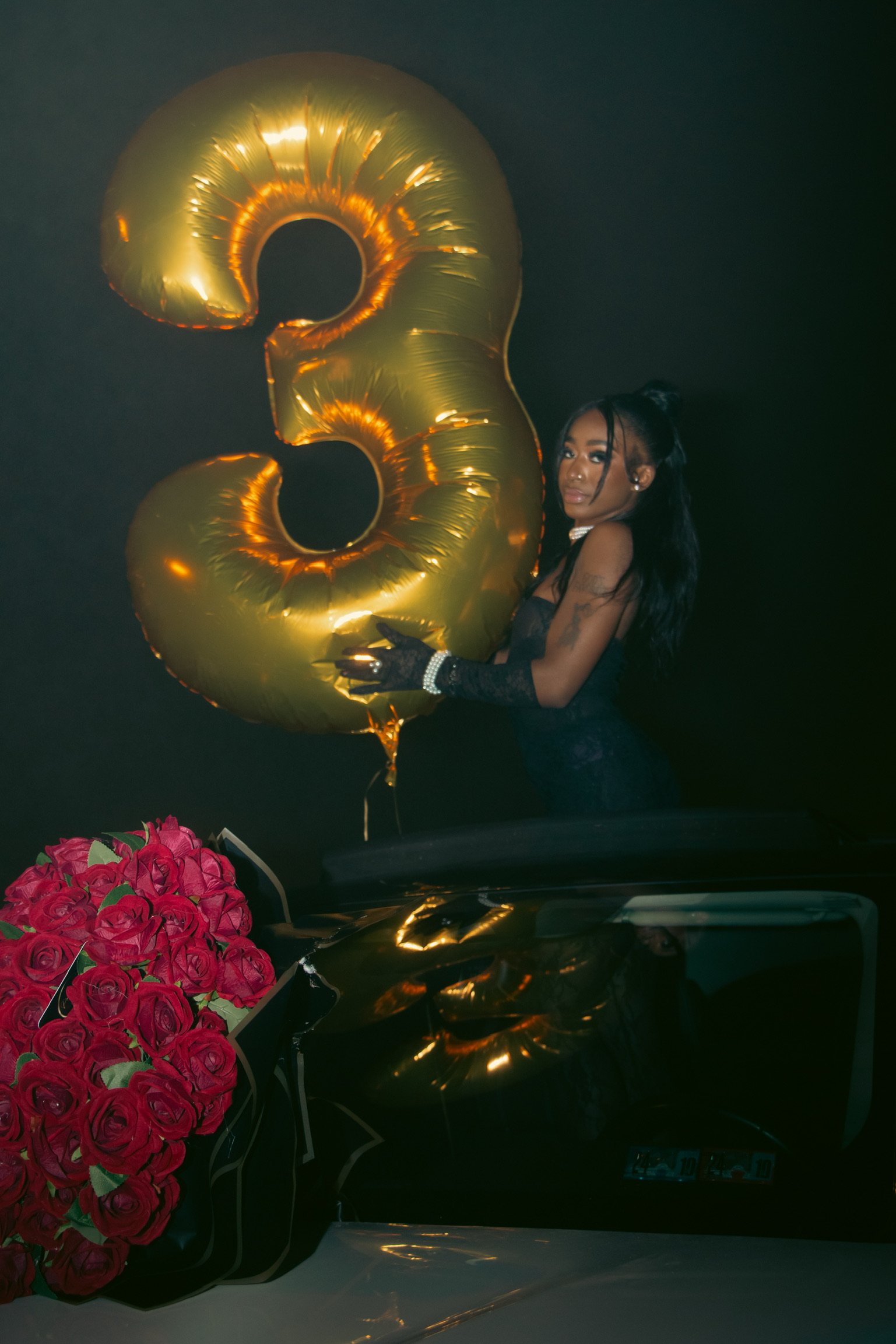 A woman in a black dress and lace gloves holding a large gold number 3 balloon, standing behind a bouquet of red roses and a black car with a blurred dark background.