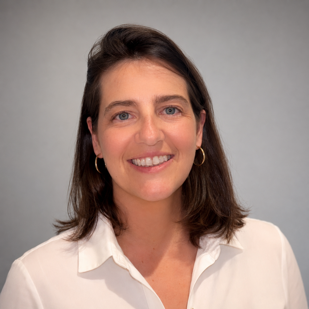 A woman with shoulder-length brown hair, wearing a white blouse and gold hoop earrings, smiling against a plain gray background.