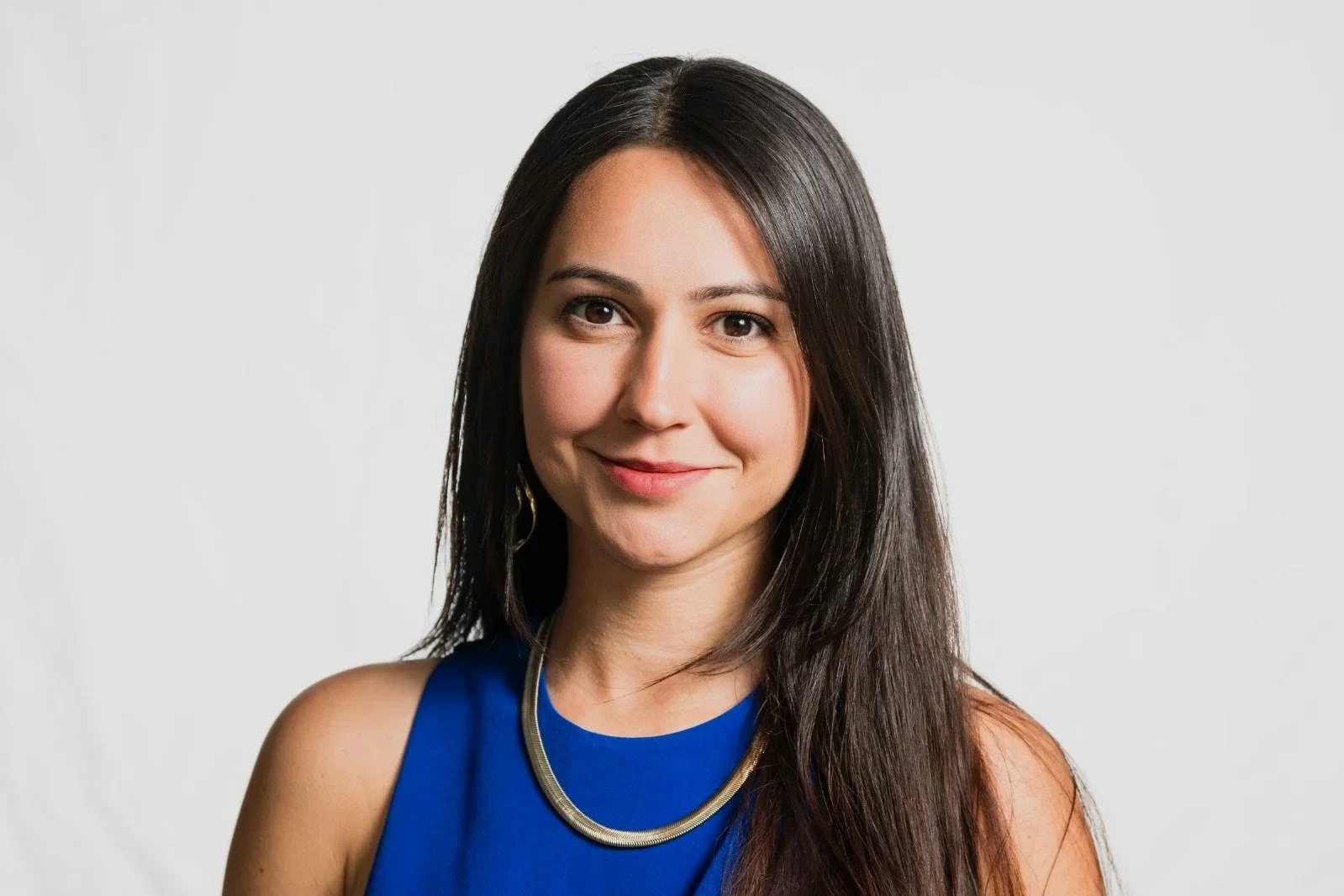 A woman with long dark hair wearing a blue sleeveless top and gold necklace, smiling against a plain white background.