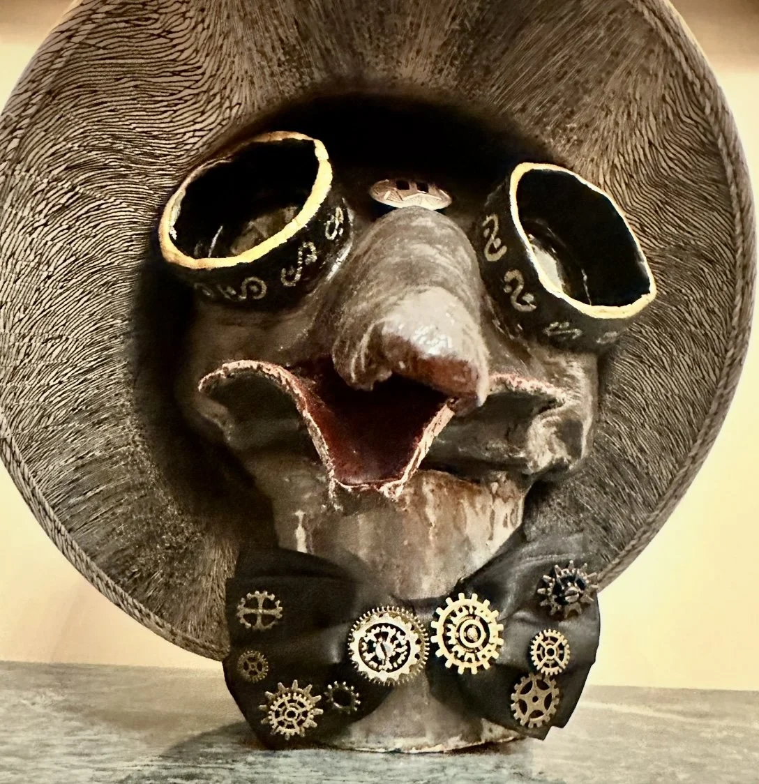 A decorative sculpture of a face with a wide-brimmed hat, goggles, a nose, an open mouth, and mechanical gears on a bowtie.