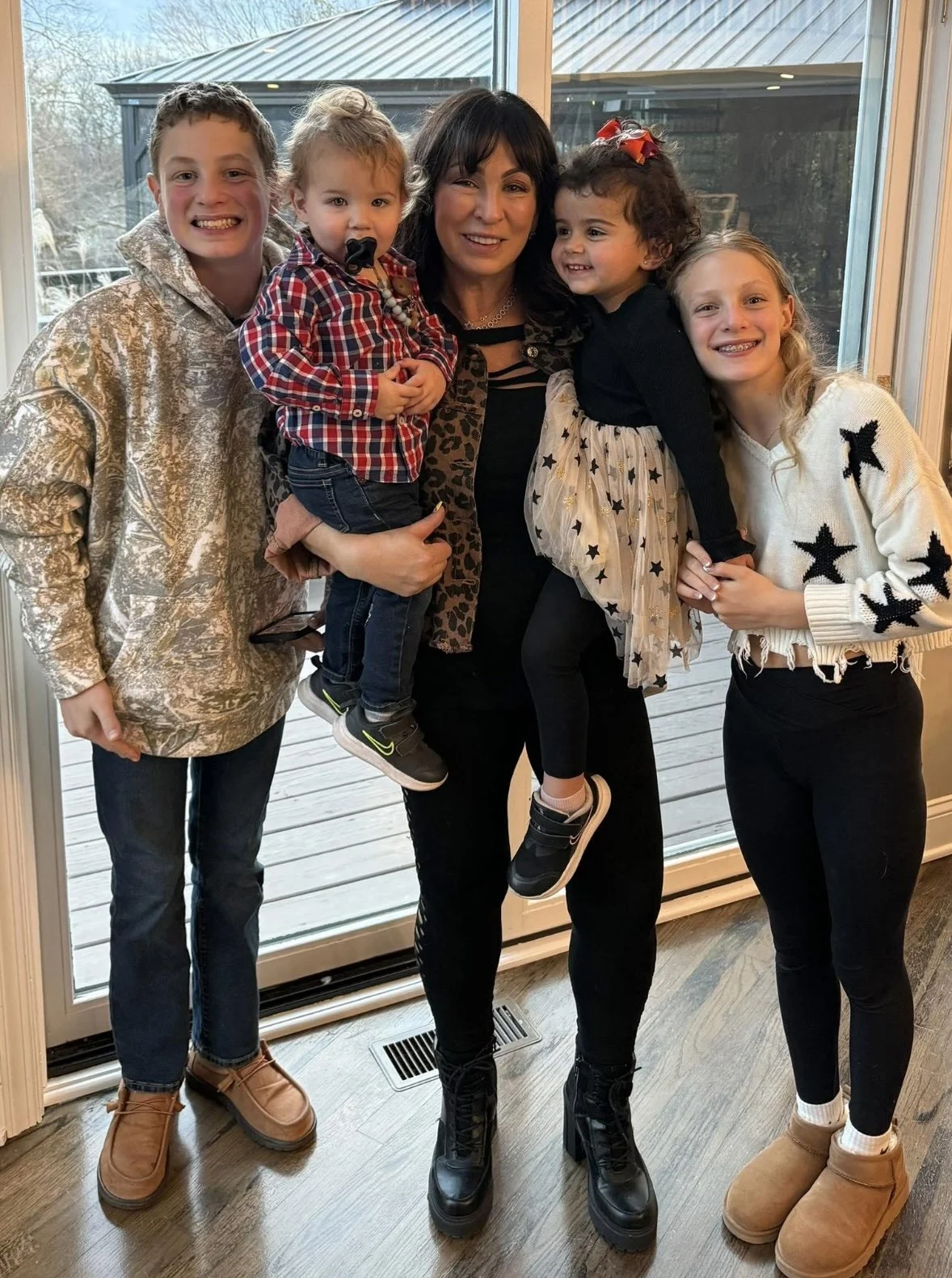 A woman with four children posing indoors by a glass sliding door, with a wooden deck outside visible through the glass.