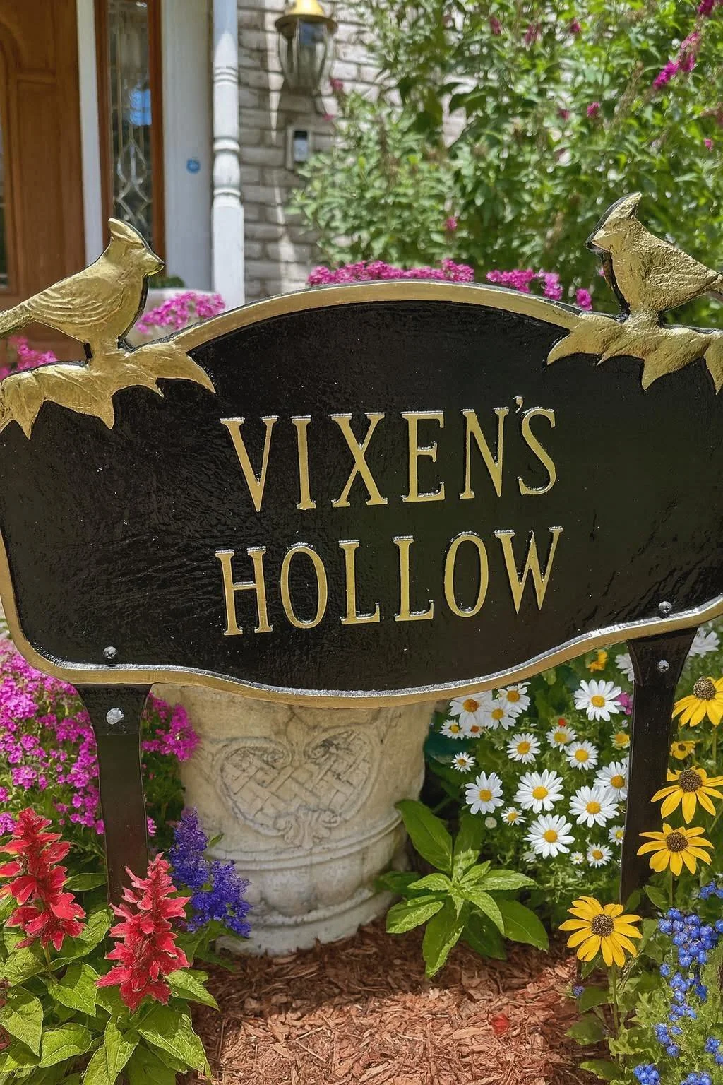 A black and gold sign with the words 'Vixens Hollow' in metal letters, decorated with gold birds on top, surrounded by colorful flowers in a garden setting.