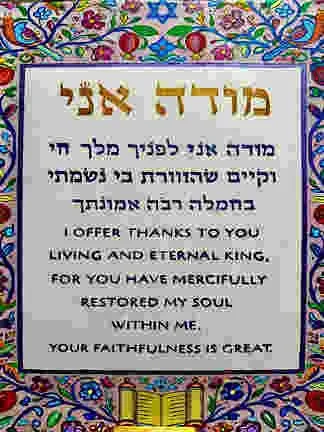 Colorful embroidered fabric with a central prayer in Hebrew and English expressing gratitude and praise to God, surrounded by decorative patterns and symbols.