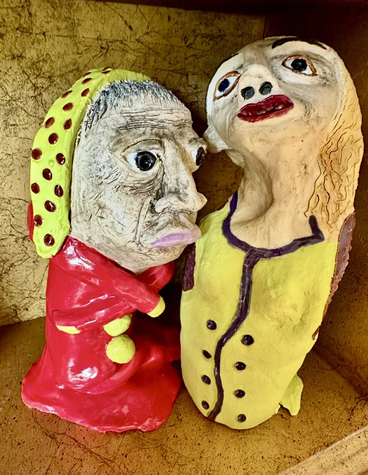 Two colorful ceramic or sculpted figures with exaggerated facial features and painted clothing, positioned close together against a brown textured background.