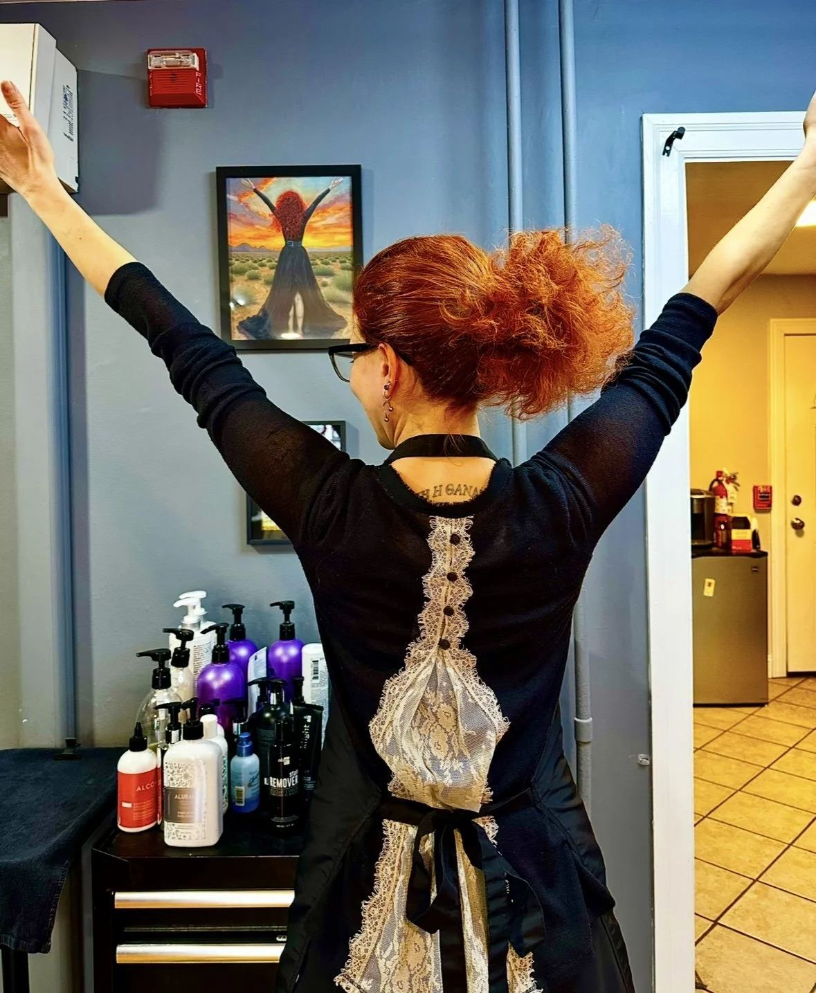 The back of a woman with her arms raised, showing her curly red hair and an apron with lace back.