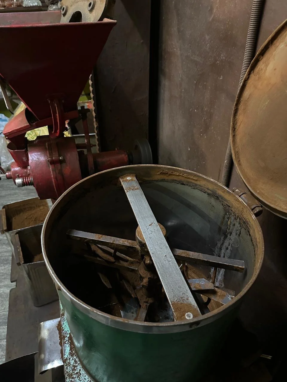 Green metal container with rusted metal blades and parts inside, next to a rusty metal surface and a red machine in a workshop.
