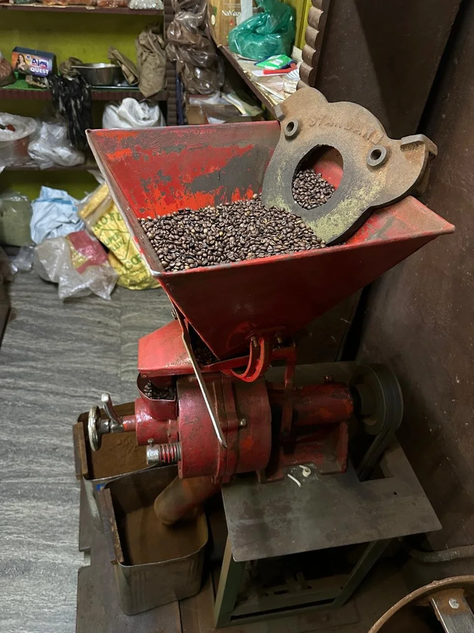 A red coffee bean grinder machine filled with coffee beans, situated in a rustic work area with various tools and supplies in the background.
