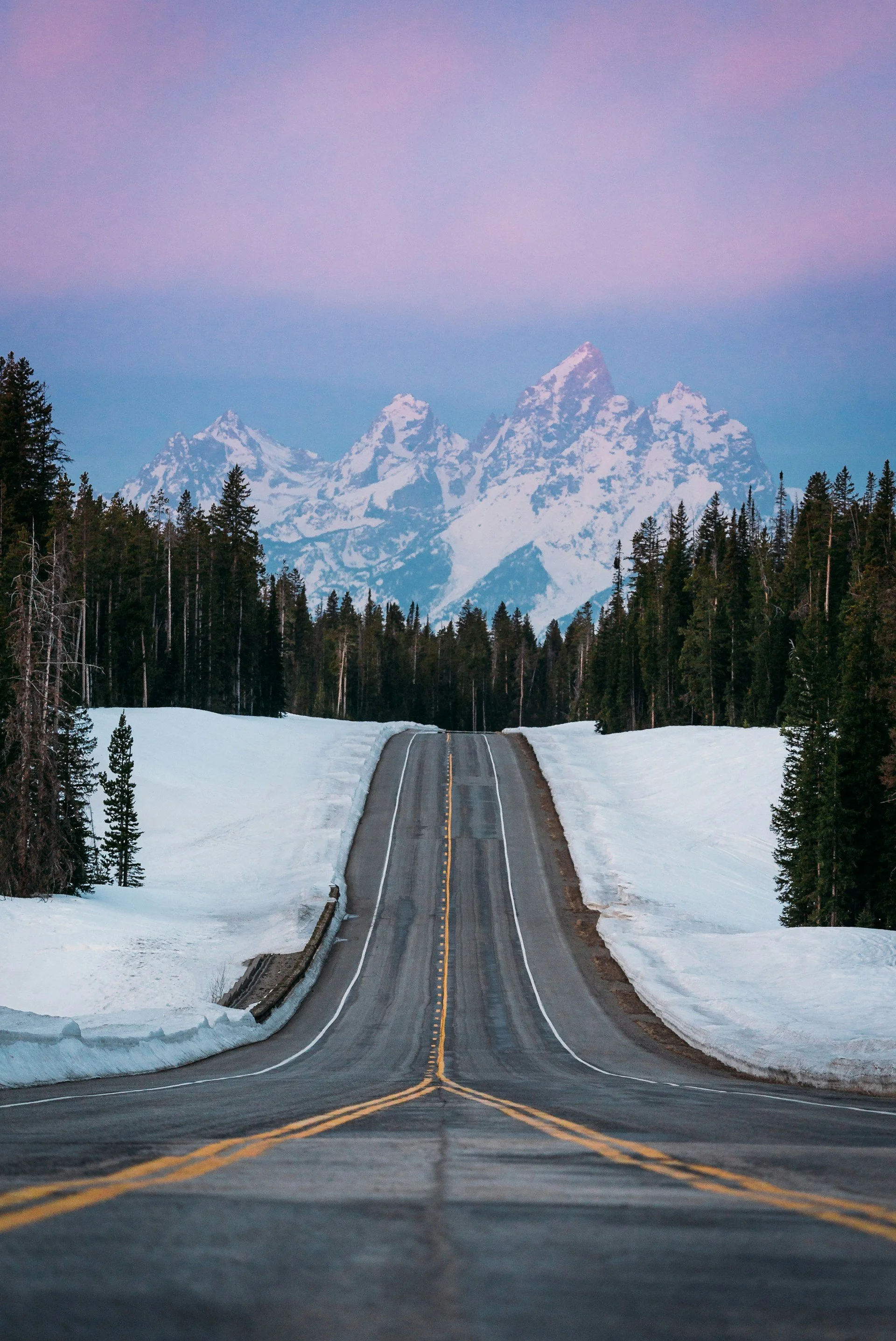 A winding road through snow-covered landscape with tall pine trees and snow-capped mountains in the background during dusk.