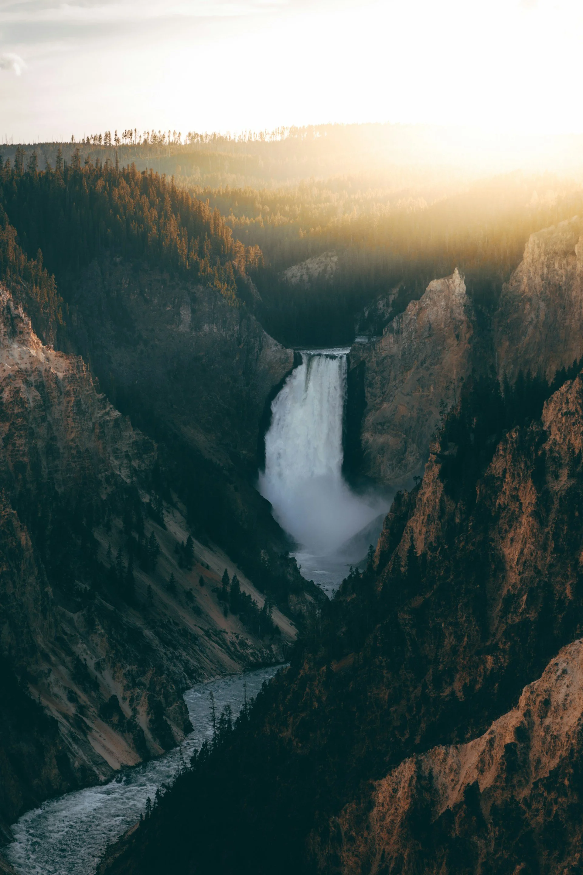 A waterfall flowing through a canyоn with steep rocky walls and a forested area at the top, illuminated by sunlight.