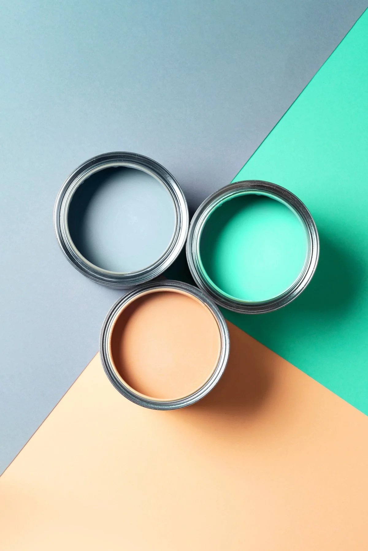 Three open cans of pastel-colored paint in blue, mint green, and peach, placed on a background of pastel-colored paper in blue, green, and peach.