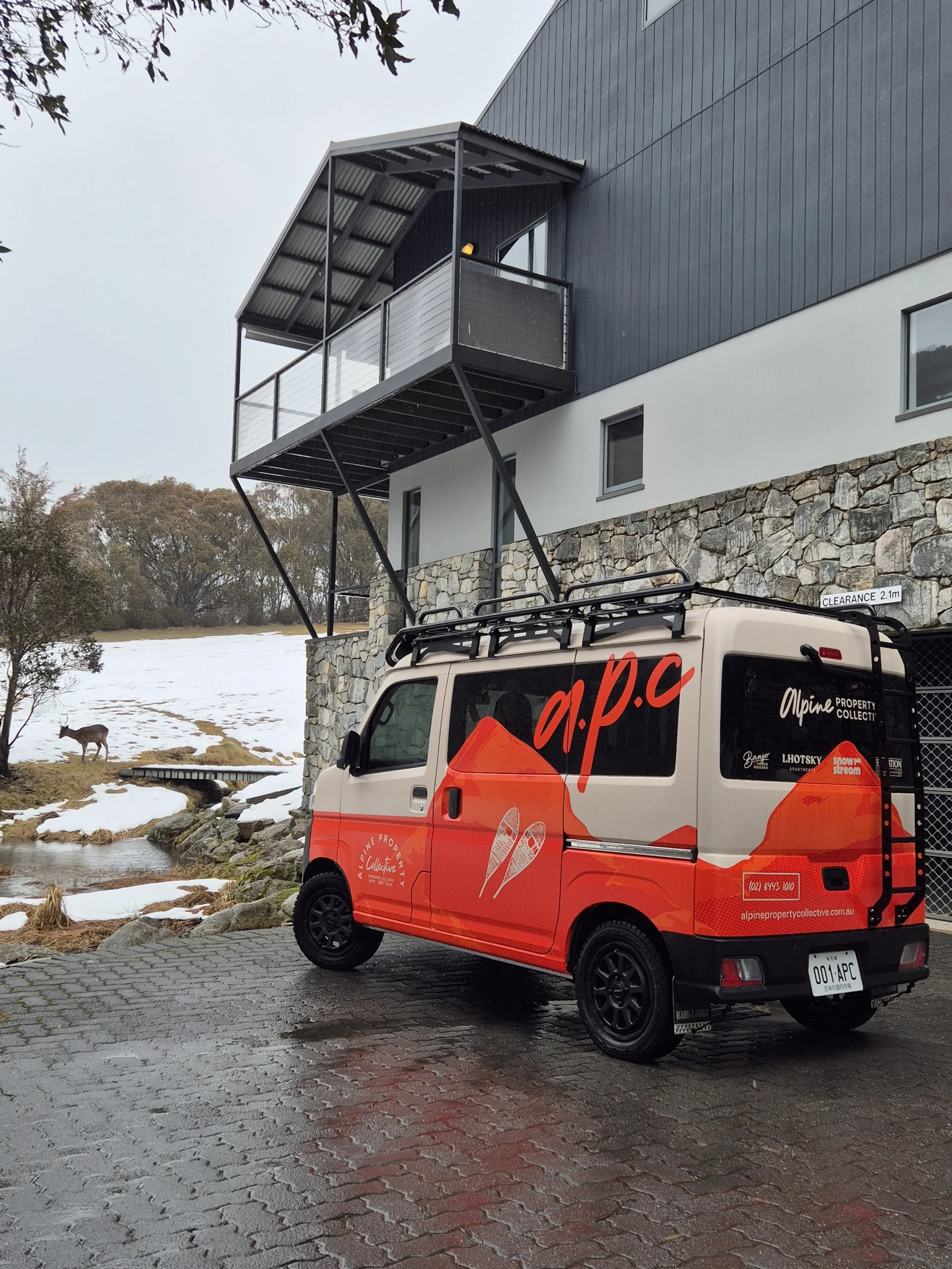 A small van with alpine property collective branding parked on a wet brick driveway outside a modern building with stone and dark siding, snowy landscape with a stream and a deer in the background.