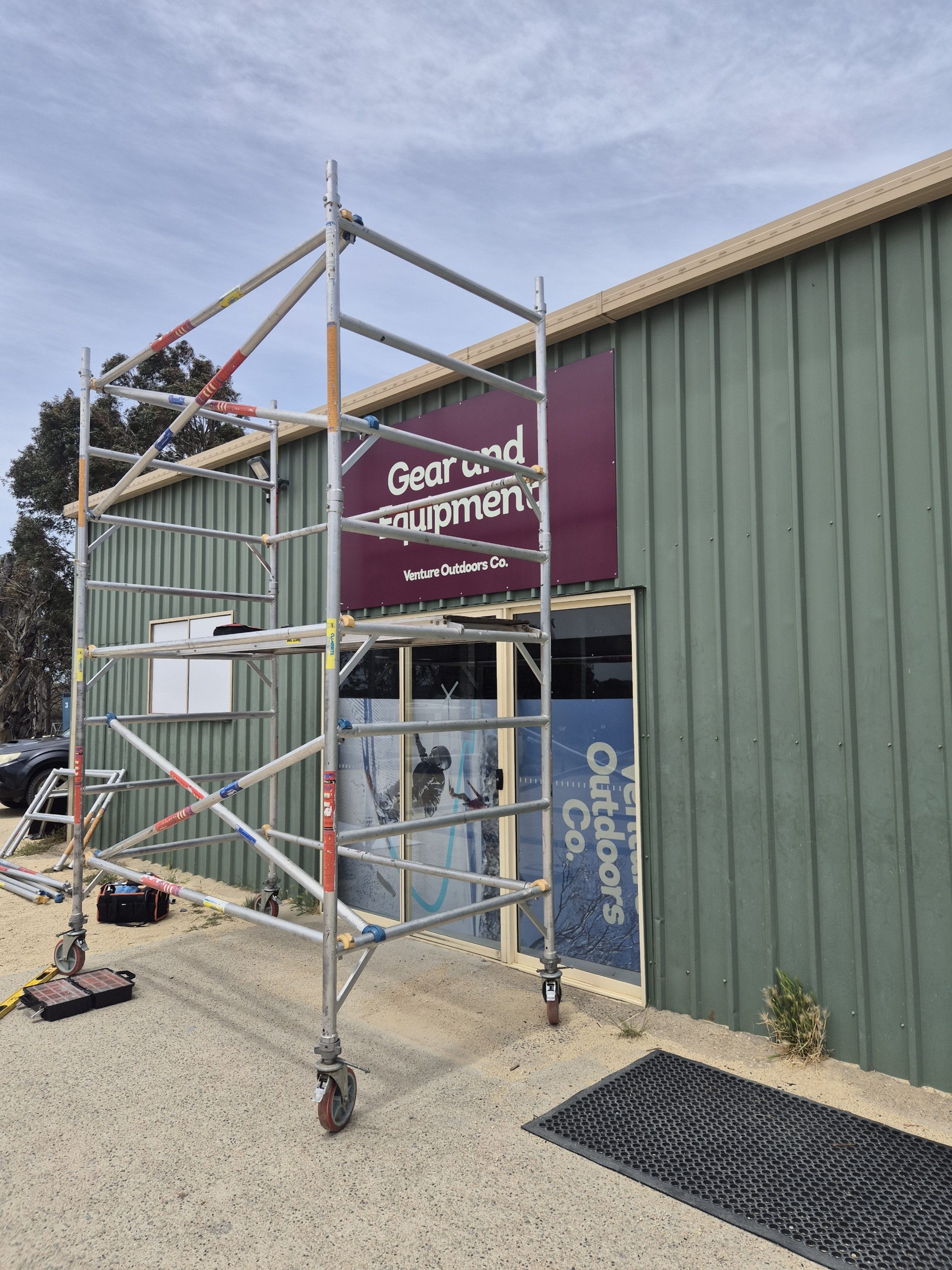 Scaffolding set up outside a green metal building with a purple sign that reads 'Gear and Equipment' and a window with 'Venture Outdoors Co.' sign.
