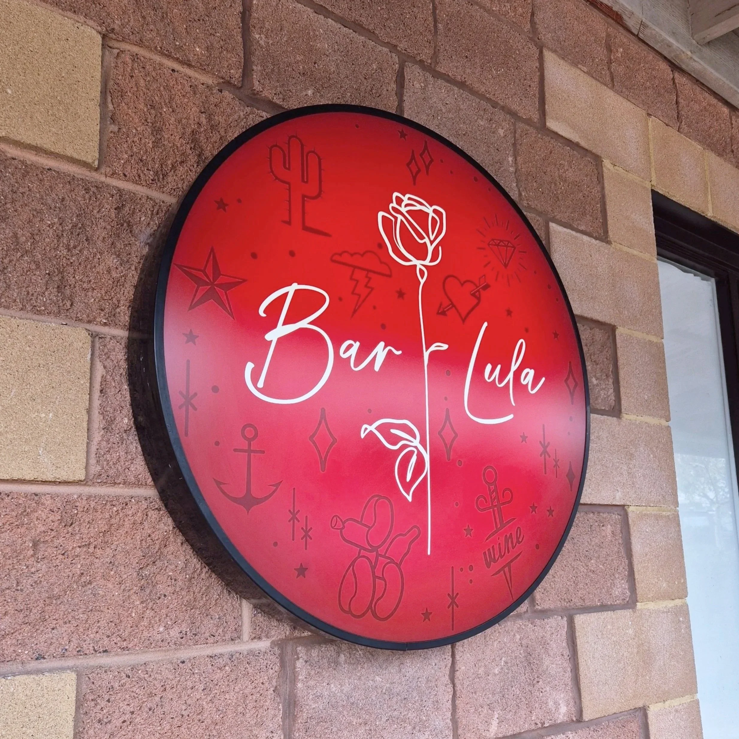 Red circular sign for Bar Lula mounted on brick wall, decorated with tattoo-style icons including a rose, anchor, hearts, star, diamond, bunny, and the words "Bar Lula" and "wine".