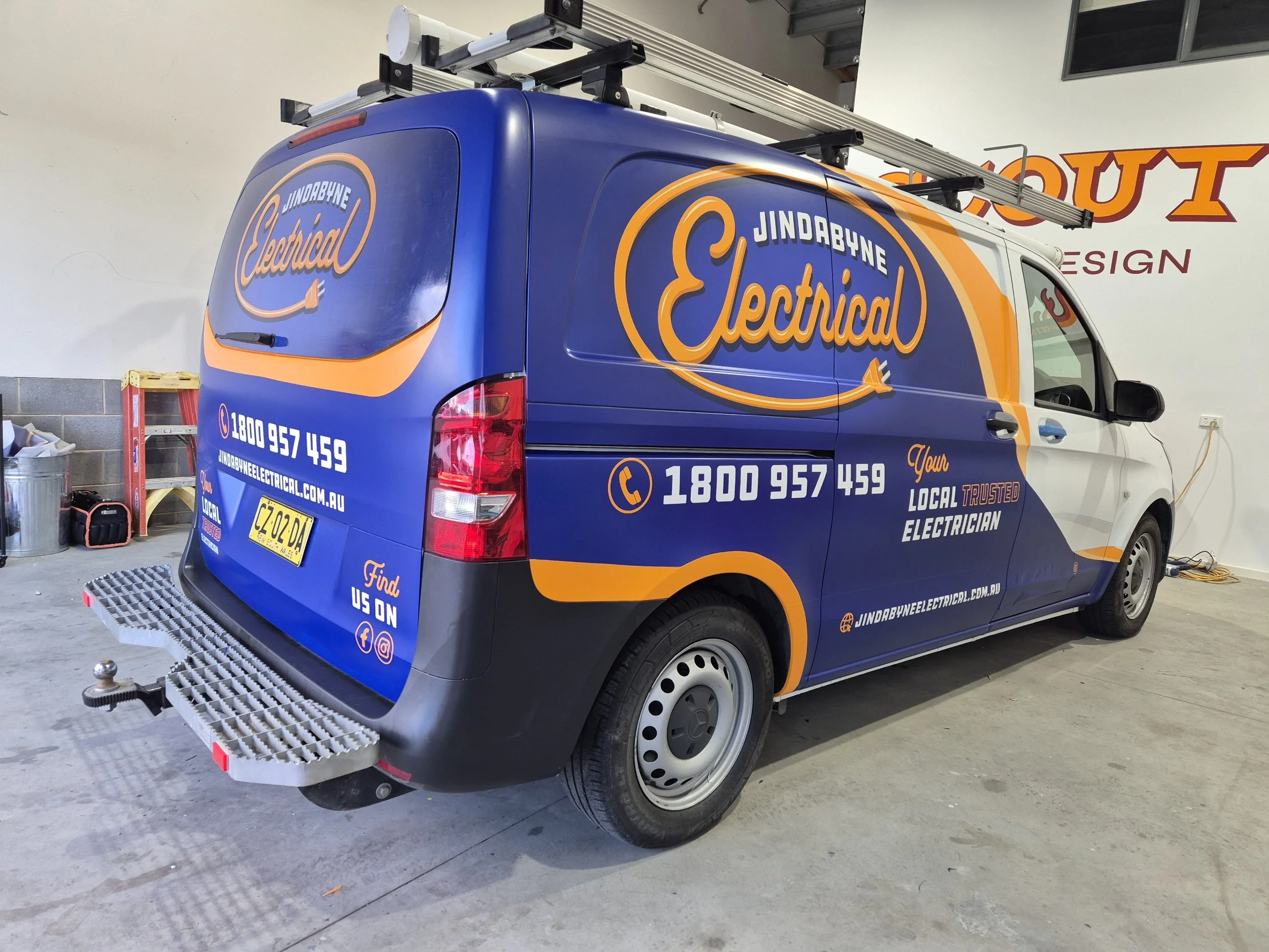 A work van with Jindabyne Electrical branding parked indoors. The van is primarily blue with orange and white accents, displaying the company's logo, contact information, website, and social media icons.