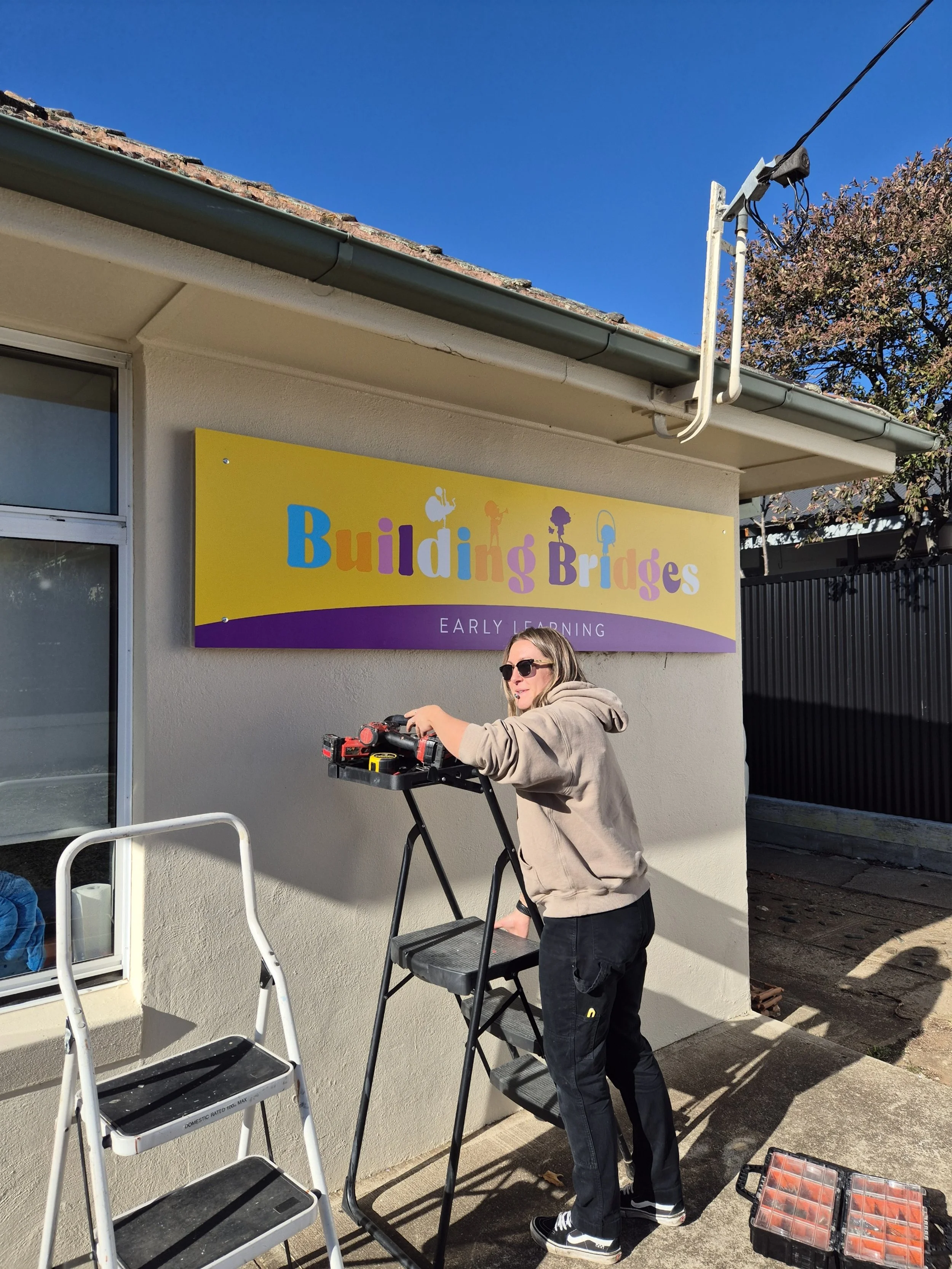 A woman with sunglasses and a beige hoodie putting up a colorful sign that says 'Building Bridges' outside a building with a sign for early learning.
