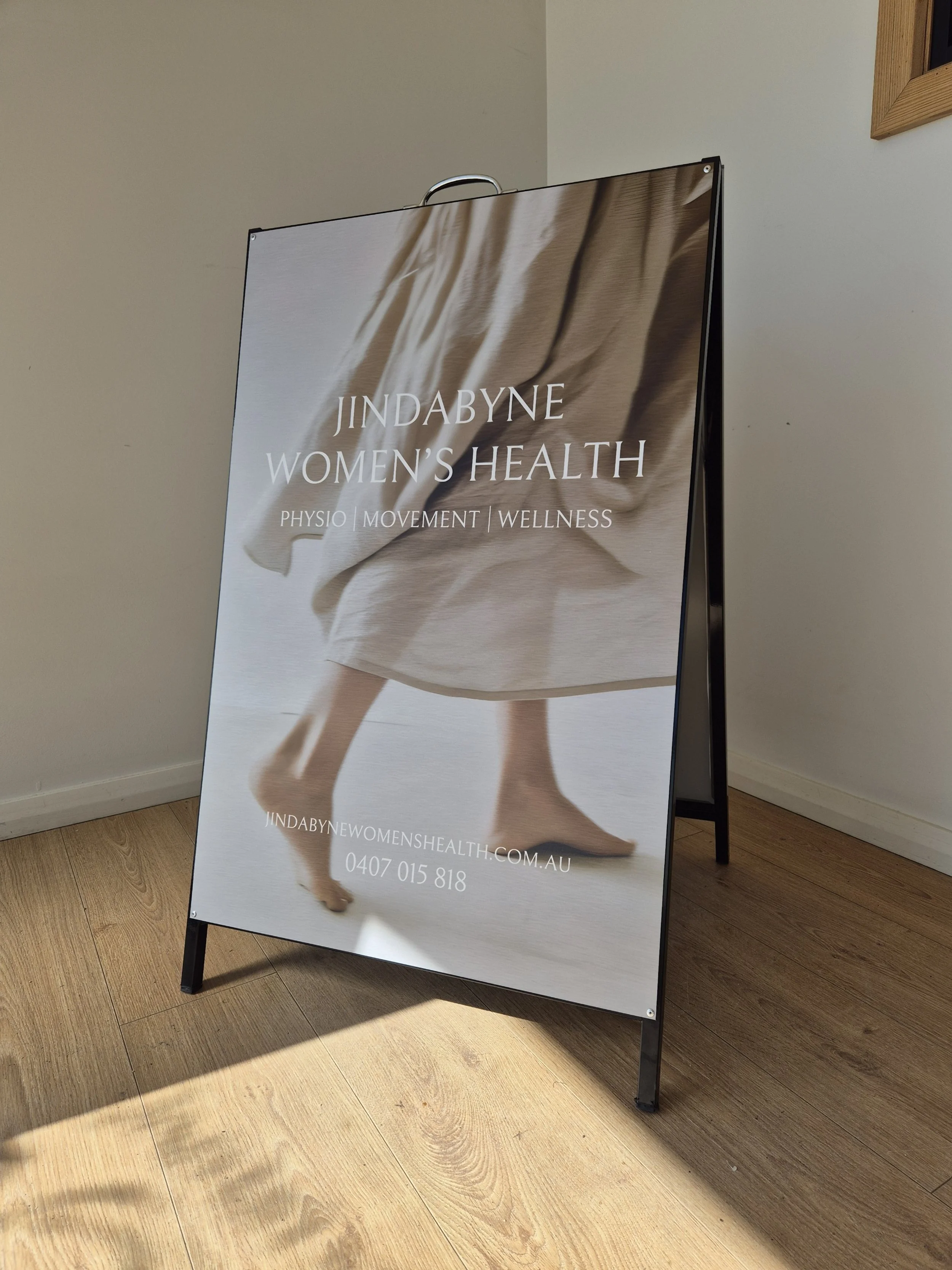 A standing signboard advertising Jindabyne Women's Health, featuring a background image of a person walking barefoot in a flowing dress. The text highlights services like physio, movement, and wellness, along with a website and phone number.