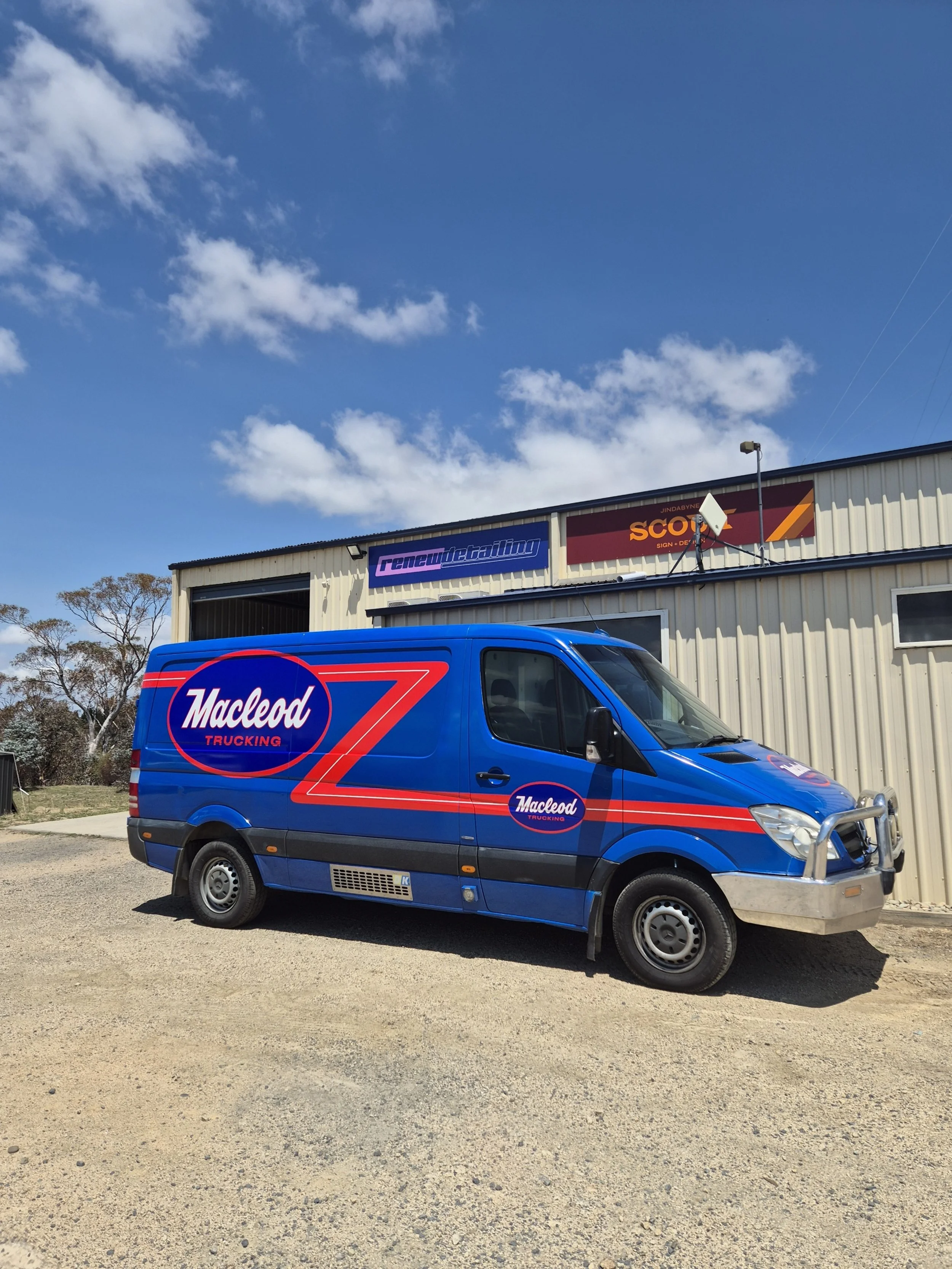 Blue Mackled trucking van parked outside a warehouse with signs for 'renew detailng' and 'scotch sign'. Clear sky with a few clouds.