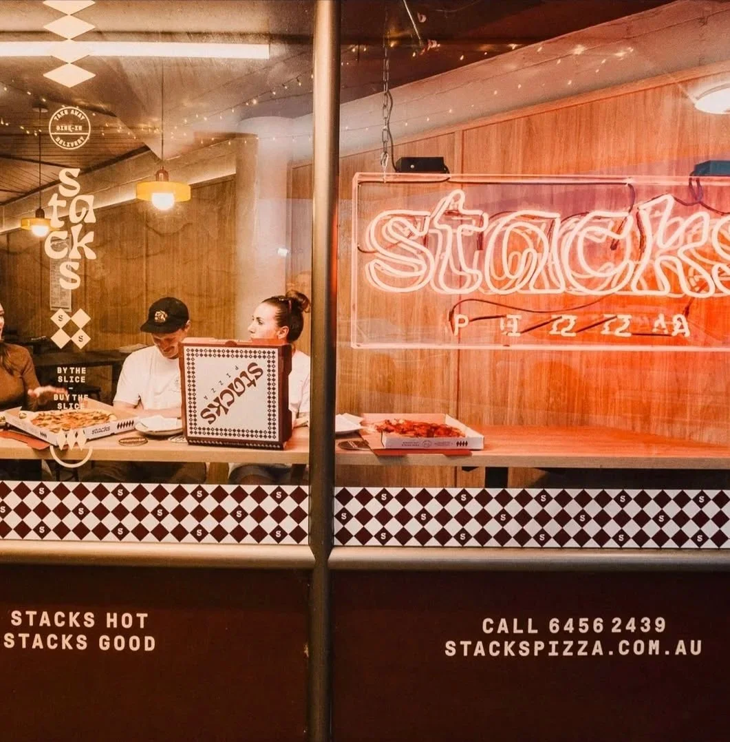 View through a restaurant window showing two women and a man sitting at a table with pizza boxes. The restaurant has warm lighting, wooden walls, a neon sign that says 'STACKS', and decorative string lights. The exterior signage advertises STACKS Pizza and includes a phone number and website.