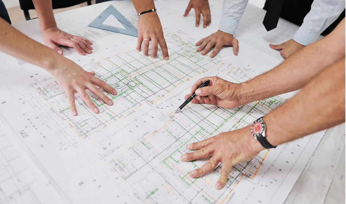 Architects and engineers discussing a detailed building plan on a large blueprint, with drafting tools on the table.