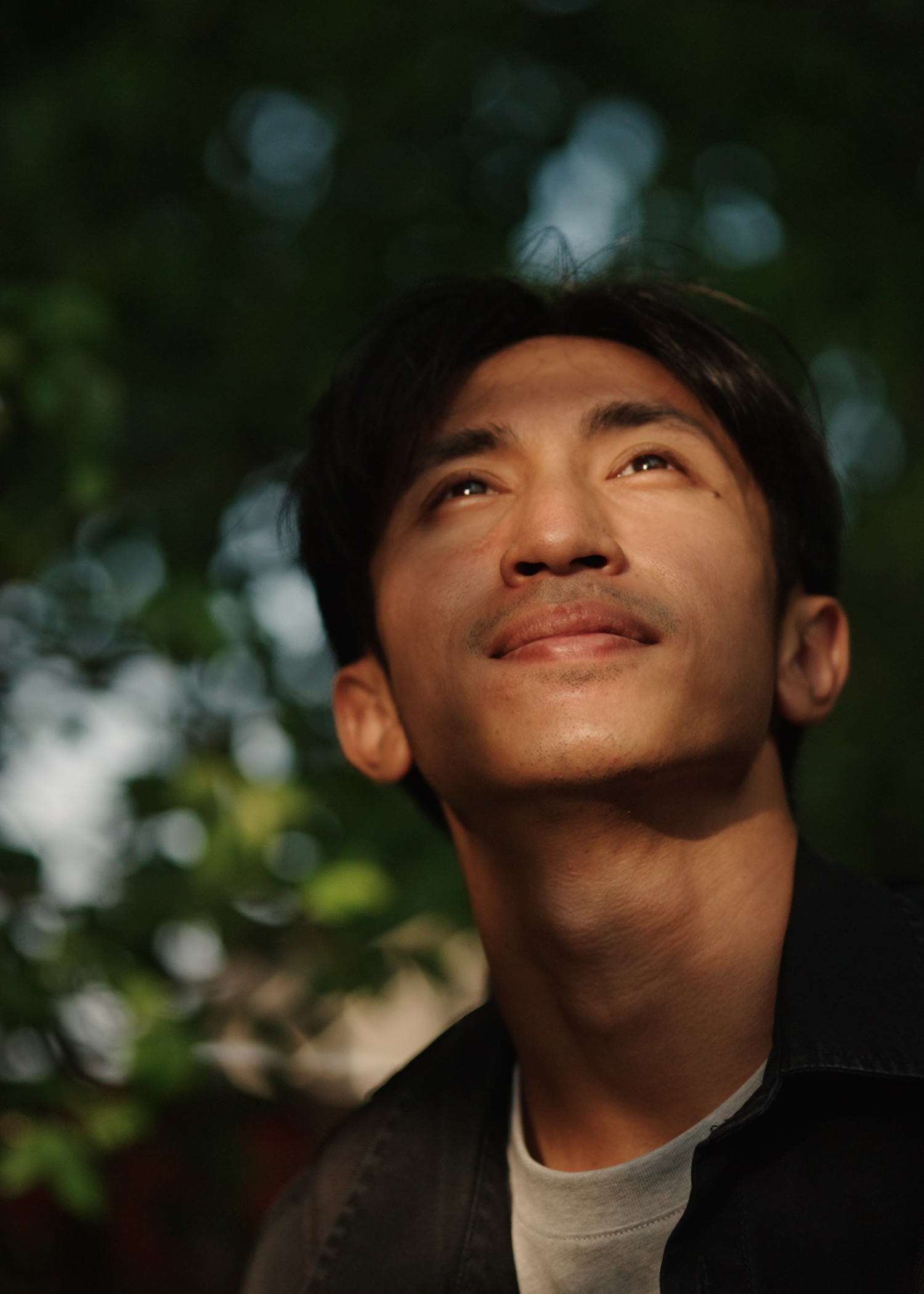 A man with black hair looking up with a slight smile, outdoors with greenery and sunlight in the background.