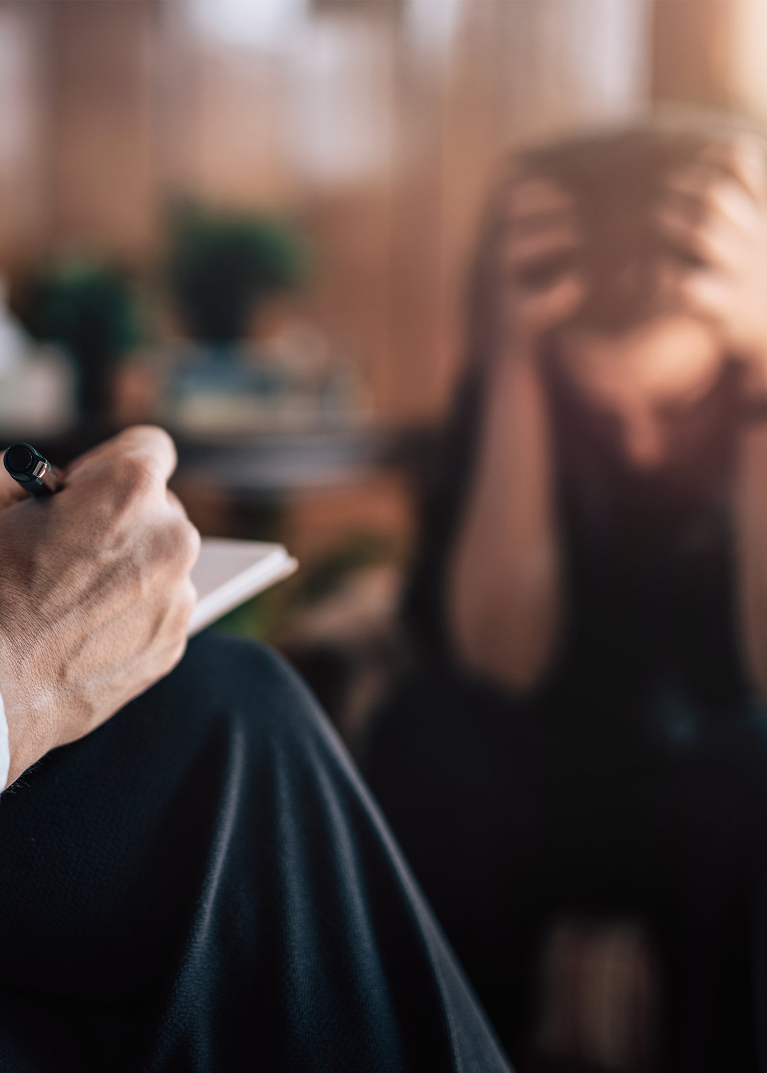 Close-up of a person's hand holding a pen and writing on a notepad, with a blurred background of a woman sitting with her face resting on her hand.