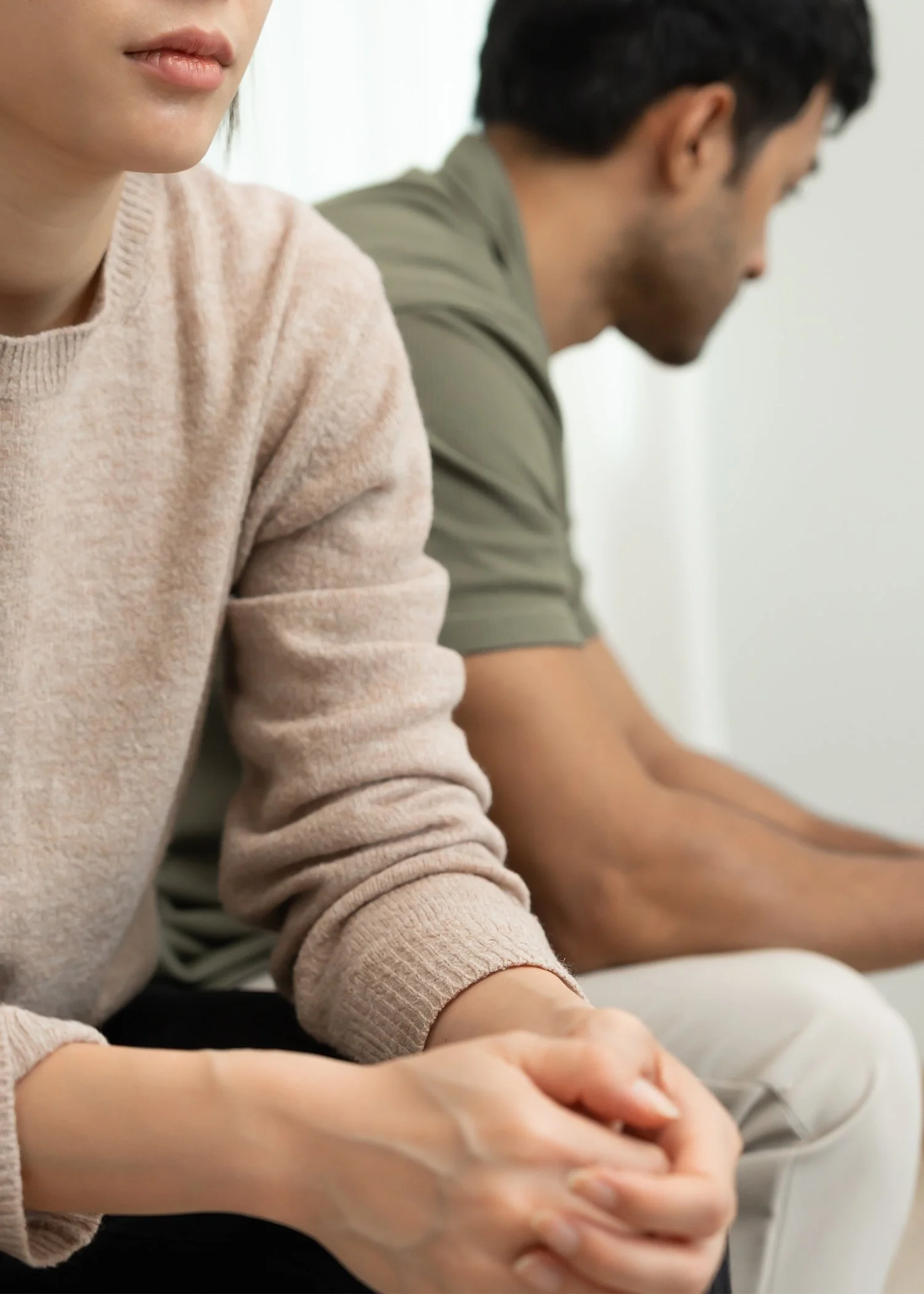An image showing two people sitting next to each other, with only their side profiles visible. The person in the foreground is a woman wearing a beige sweater, with her hands clasped and resting on her lap. The person in the background is a man wearing a green shirt, looking down.
