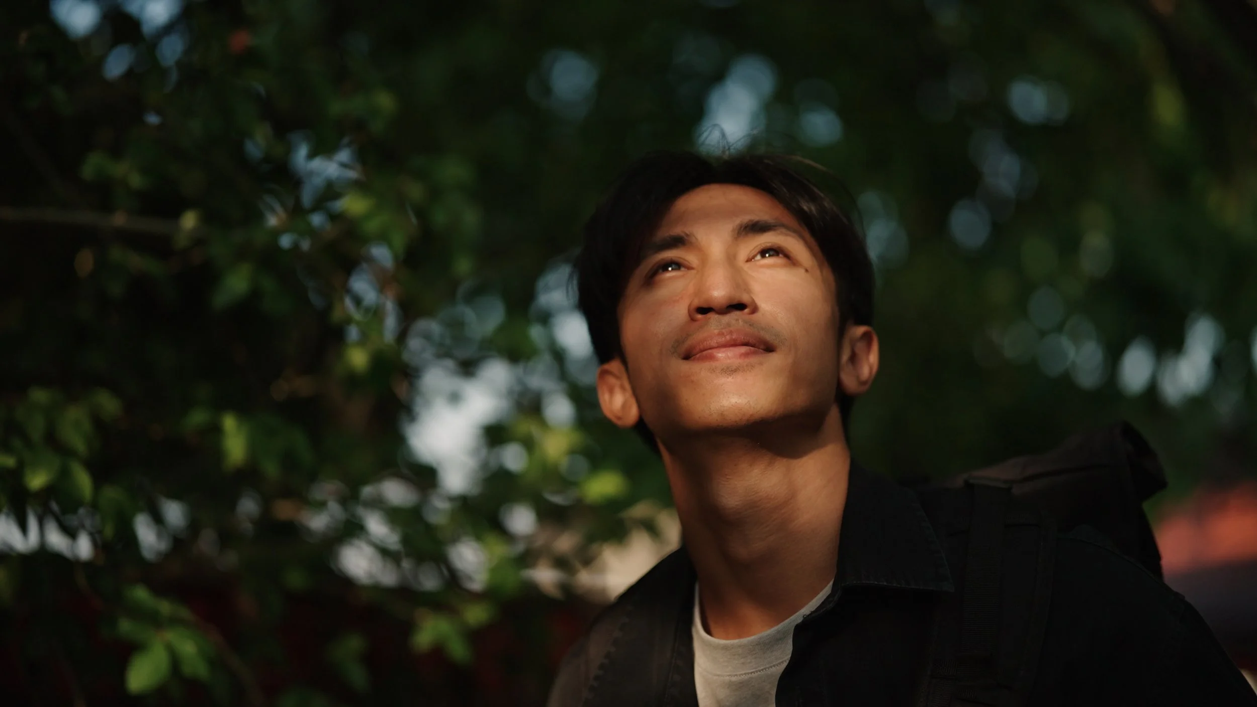 A young man with dark hair and a light complexion looking upwards with a gentle smile, outdoors with green foliage and bokeh lights in the background.