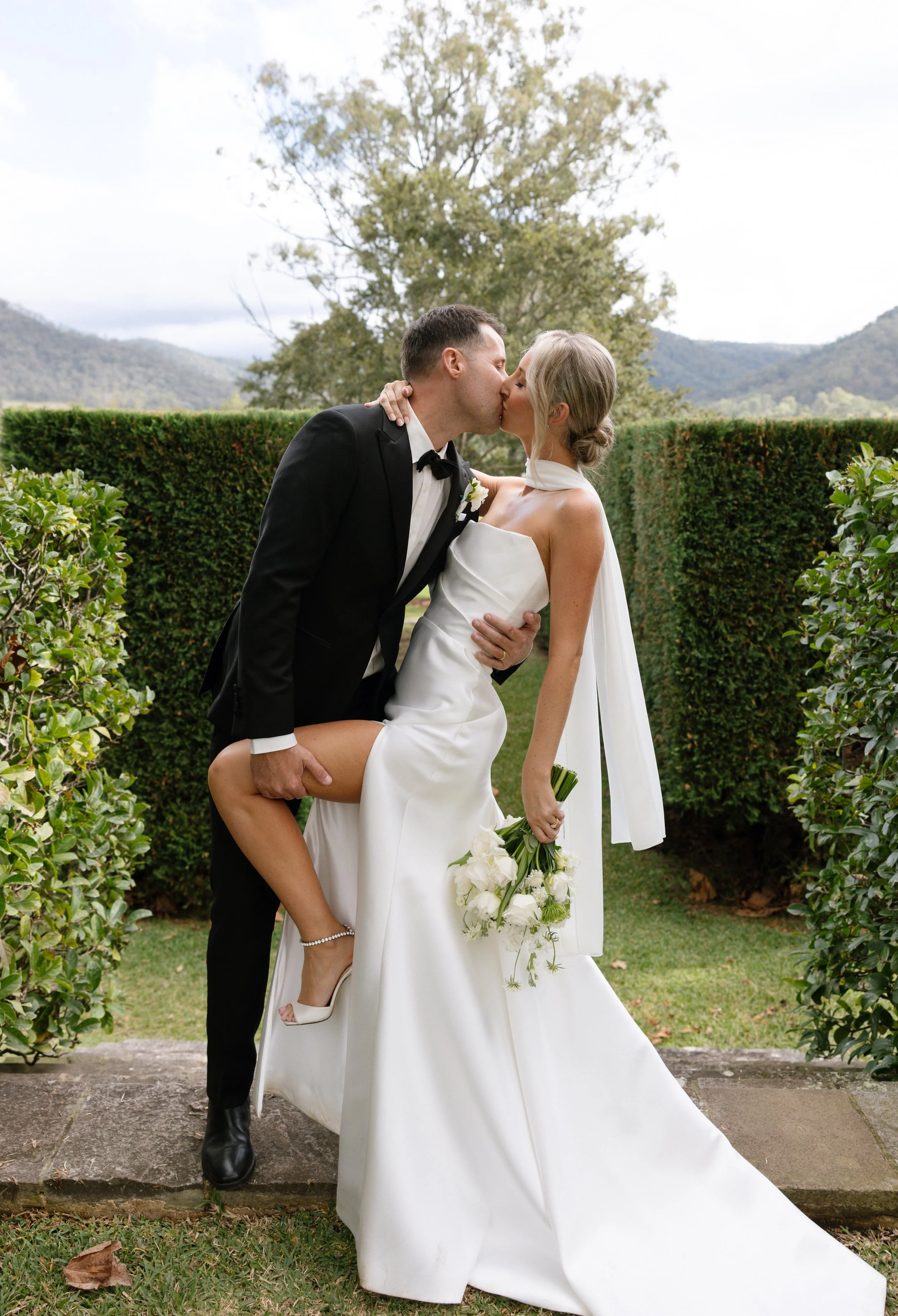 A newlywed couple sharing a kiss outdoors, the groom in a black tuxedo and the bride in a white wedding gown, with the bride holding a bouquet of white flowers.