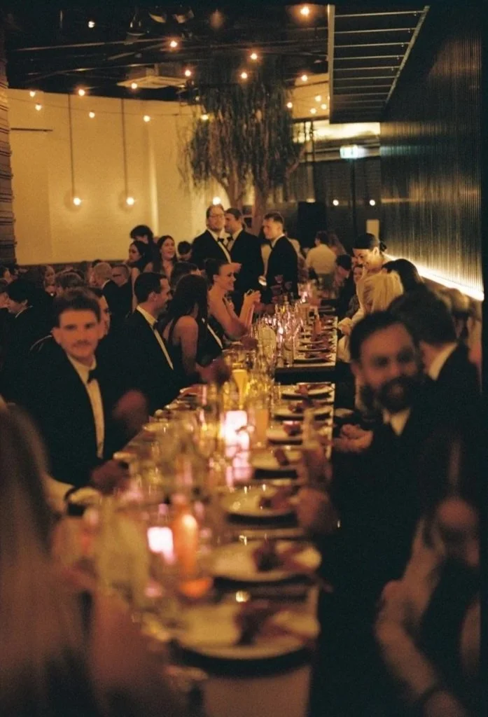 People dressed in formal black-tie attire sitting at a long banquet table at a fancy event or wedding reception, with dim lighting and hanging Edison bulbs, in a decorated indoor setting.