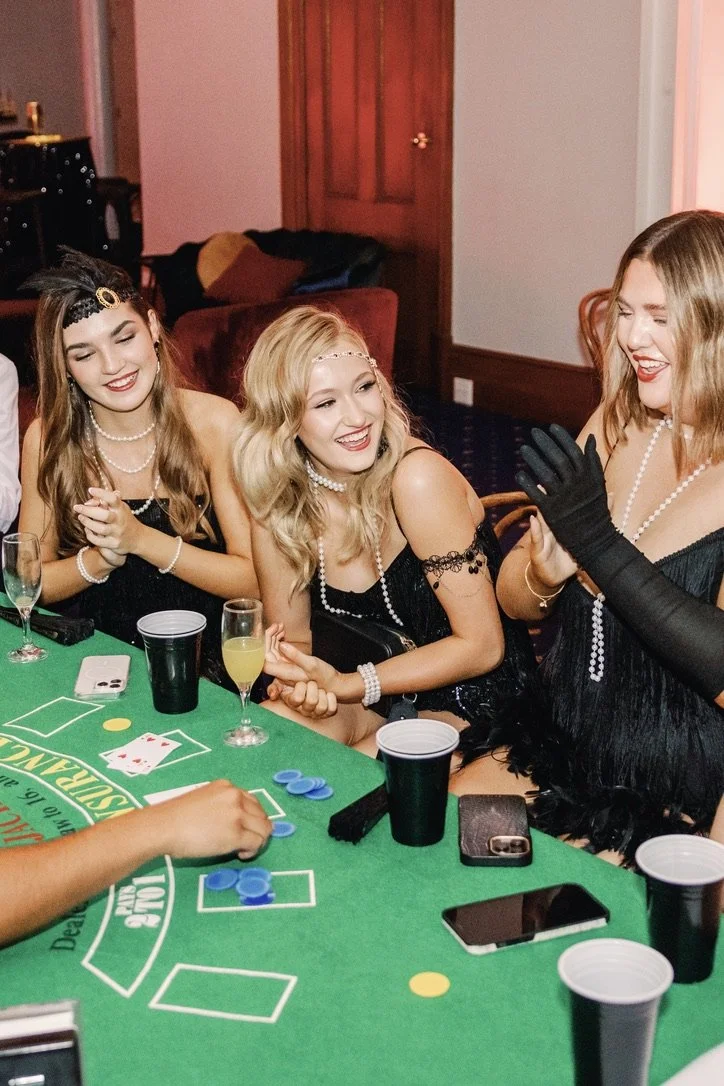 Three women in black dresses and pearl necklaces laughing and playing a card game at a party, with drinks and phones on the table.