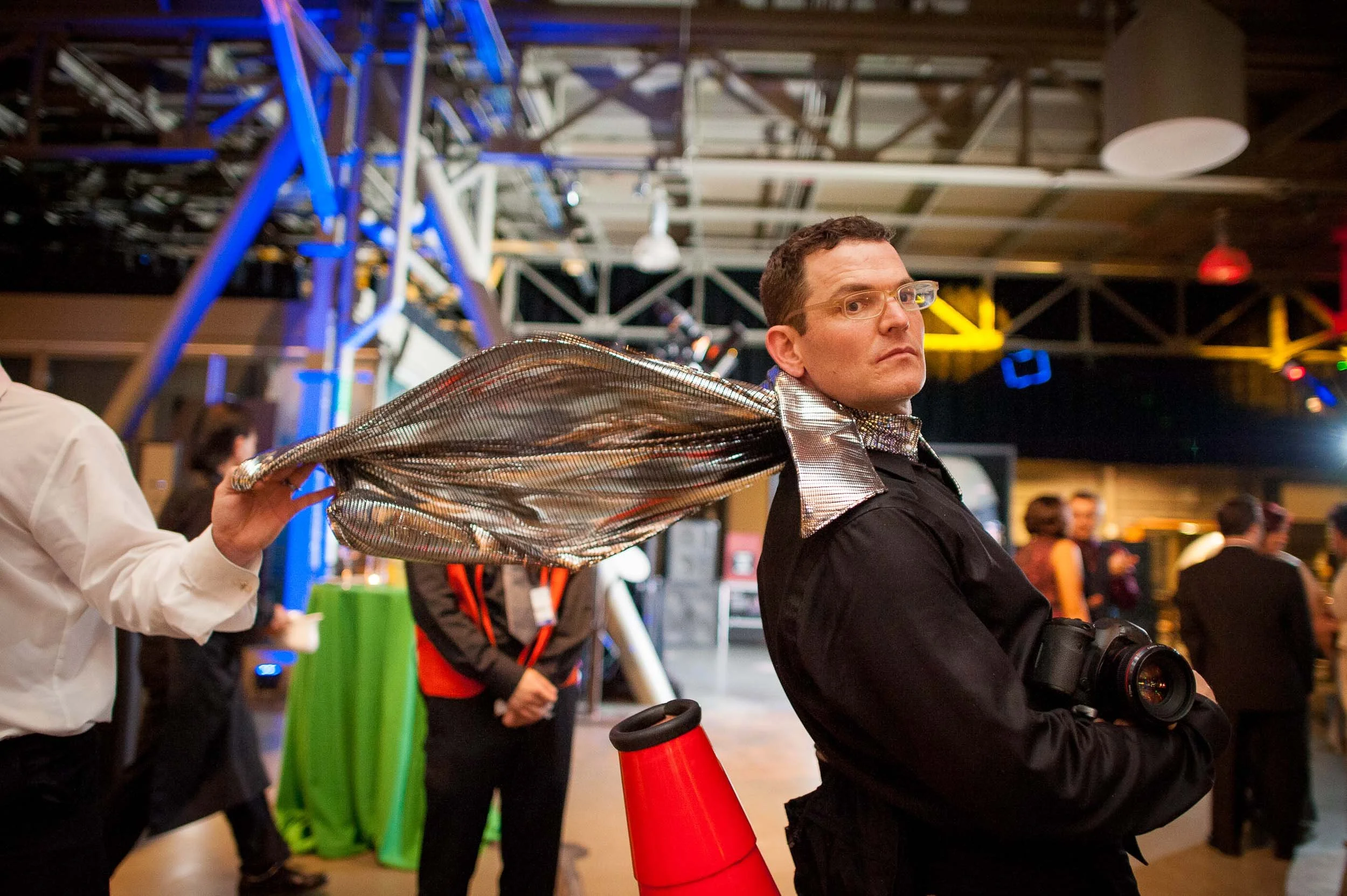 A man with glasses wearing a black outfit with a shiny silver cape around his neck, holding a camera, stands in a busy indoor event space with colorful lighting and a large metal structure in the background.