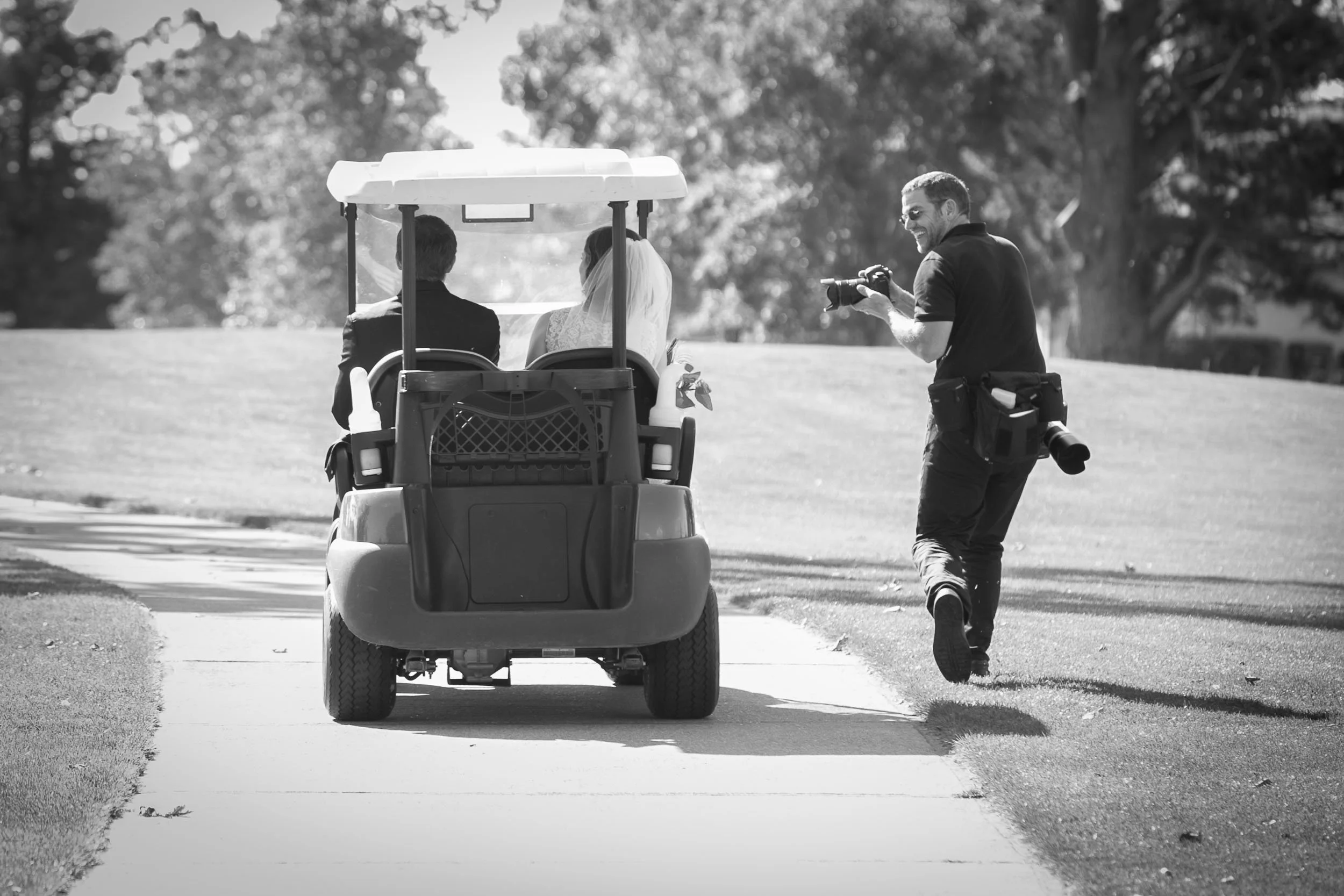 A photographer taking pictures of a bride and groom sitting in a golf cart on a golf course.