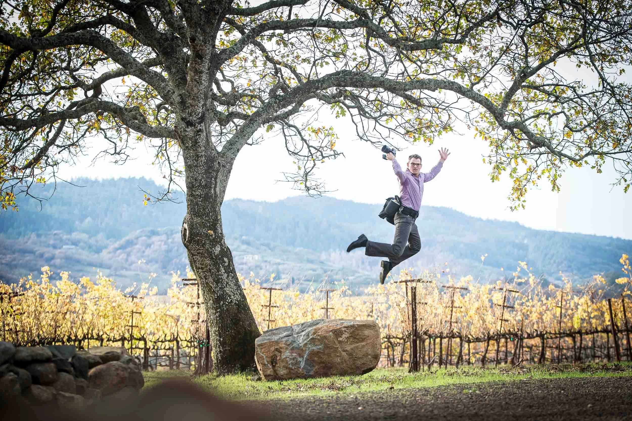 A man jumping in the air with his arms raised and holding a camera, by a tree in a vineyard with yellow autumn leaves, mountains in the background.