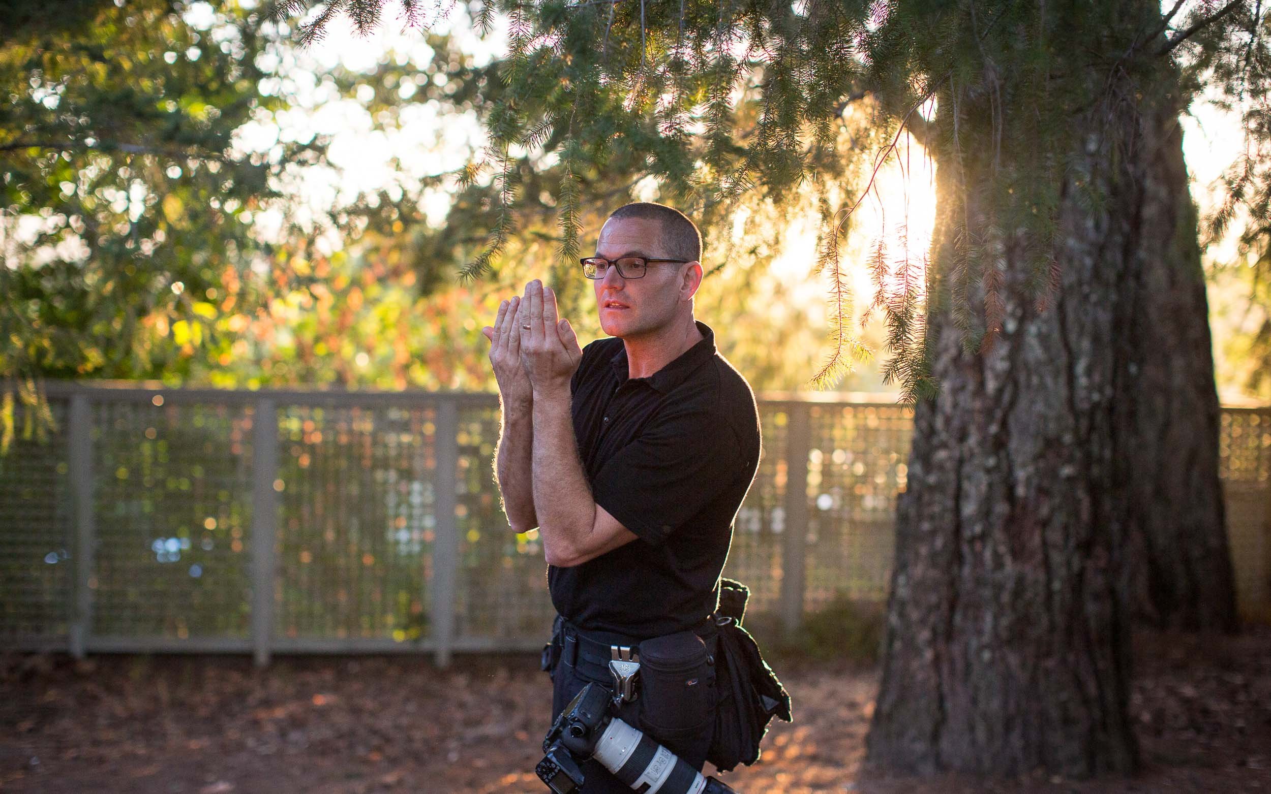 A man with glasses and a black shirt standing outdoors near a tree during sunset, holding his hands together in front of his face in a prayer-like gesture.