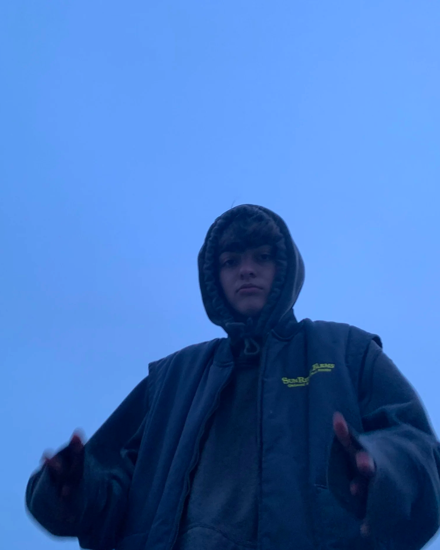 A young person wearing a hooded jacket looks down at the camera against a blue sky.