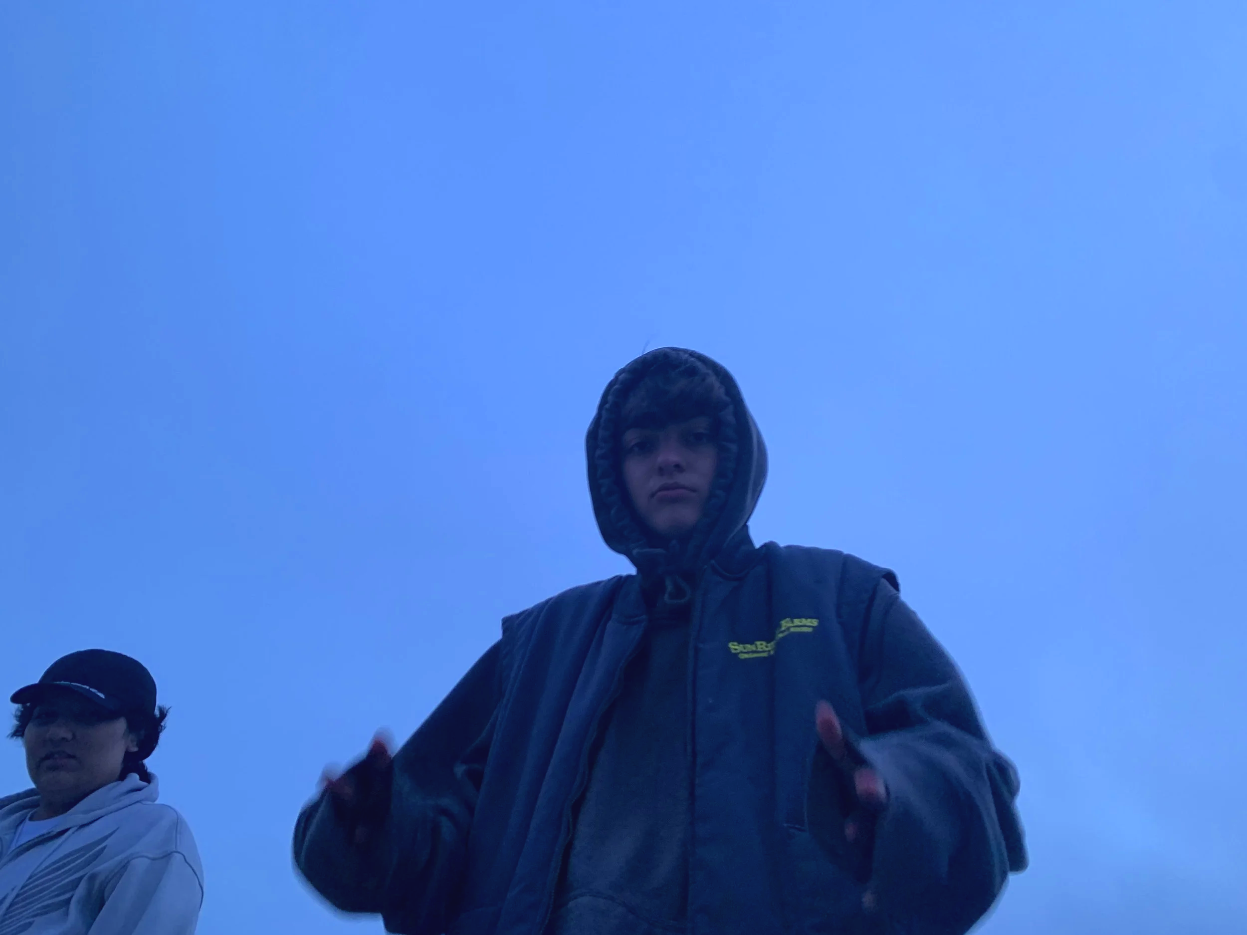 A young boy wearing a dark hoodie with the hood up and a jacket stands outdoors against a clear blue sky. Another person, wearing glasses and a hat, is partially visible on the left side of the image.