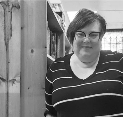 black and white image of Trudi leaning aginst a book shelf. She is wearing glasses and a black sweater with white stripes