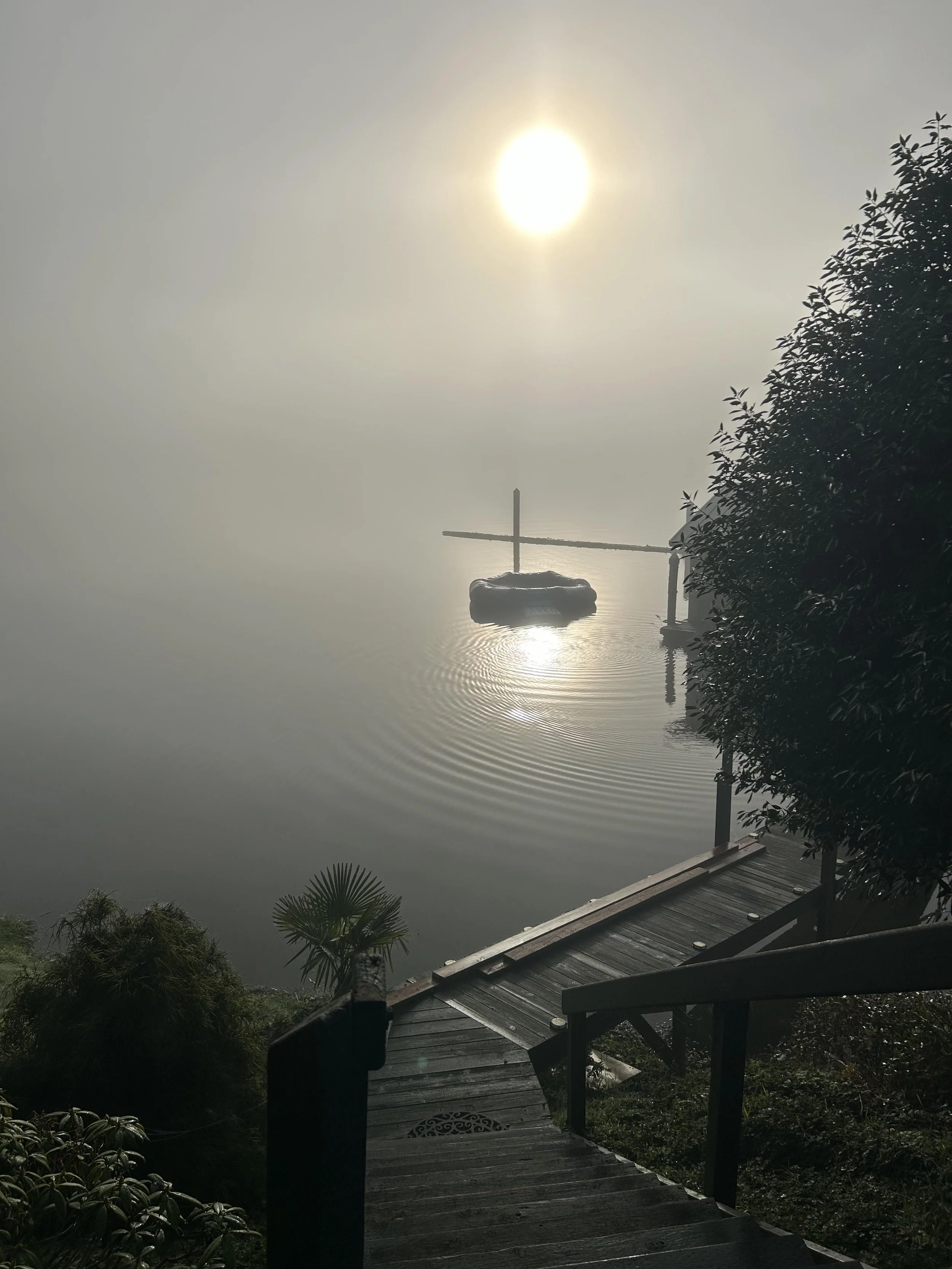 A foggy scene of a wooden dock leading down to a calm body of water with a small boat and a bamboo raft. The sun is shining through the fog, reflecting on the water, with greenery on the right side.