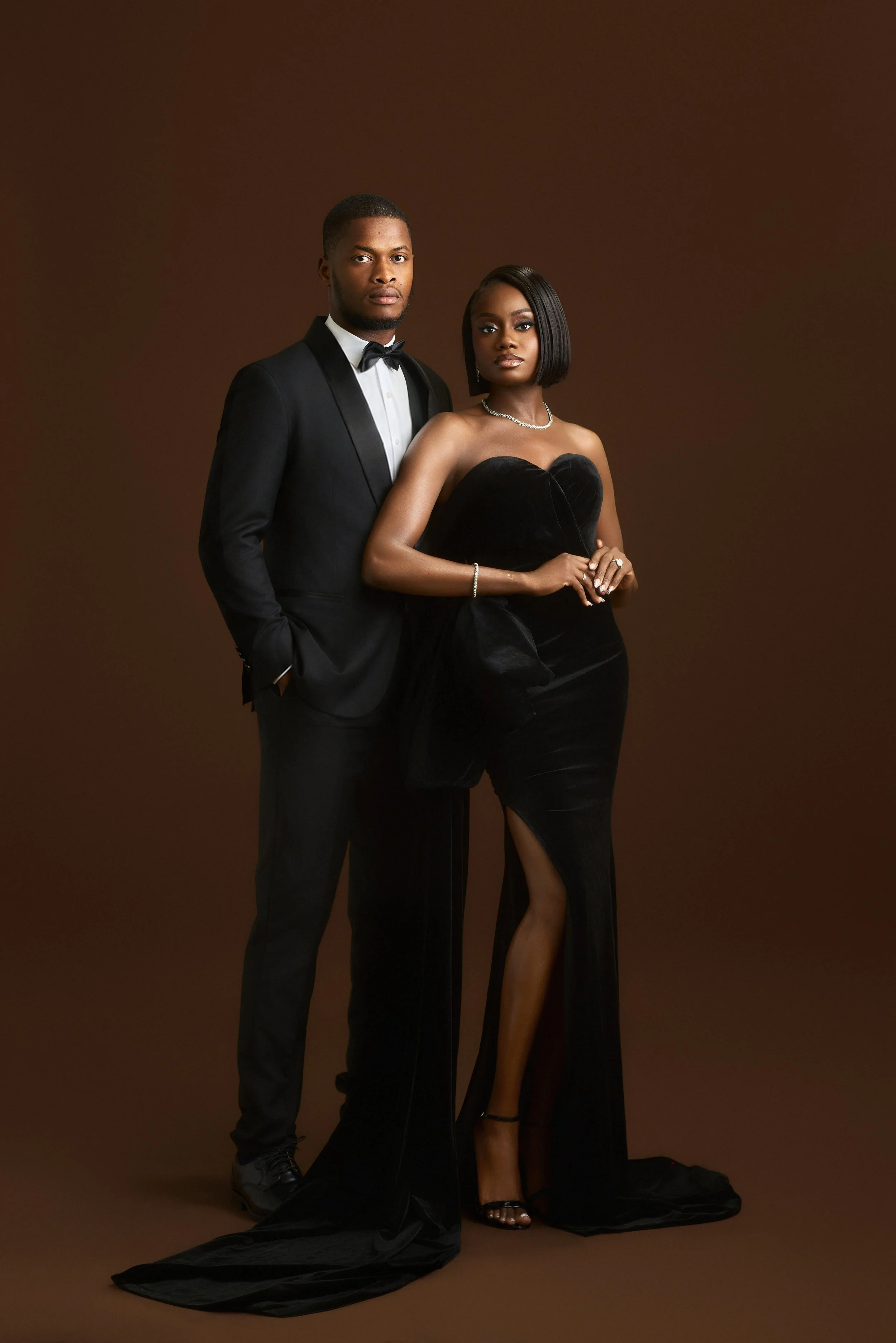 A well-dressed man in a black tuxedo and bow tie stands next to a woman in a black strapless gown with a slit, wearing jewelry and high heels, against a plain brown background.