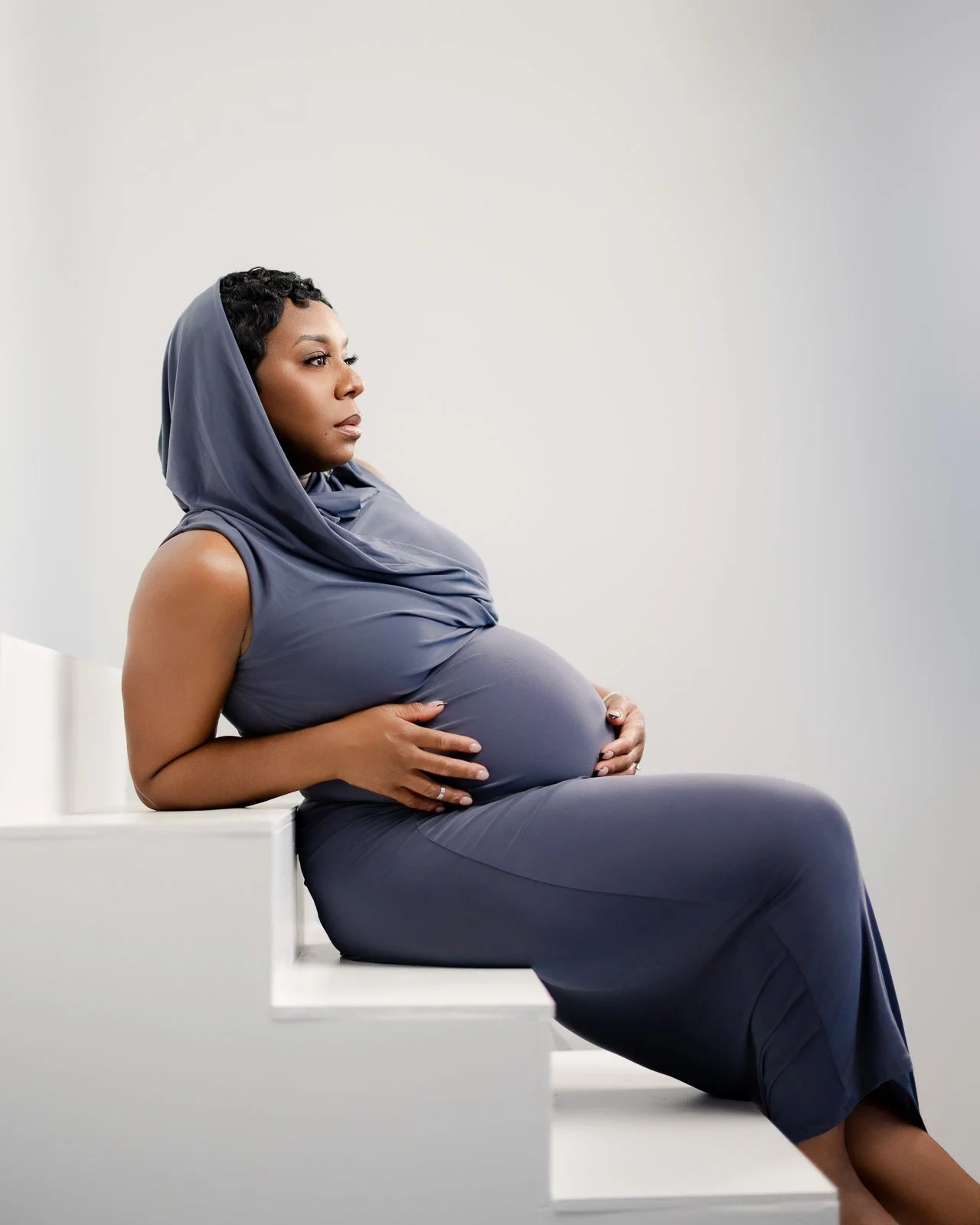 A pregnant woman in a gray dress and matching headscarf sitting on white stairs, holding her belly and looking to the left, against a plain, light-colored background.