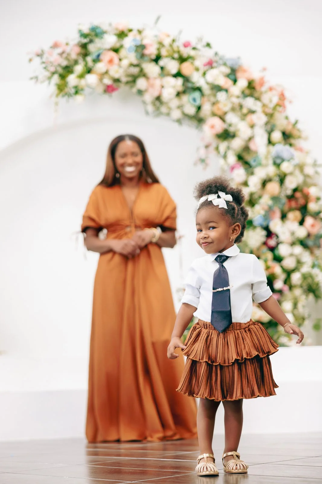 Young girl with curly hair and bow, wearing a white shirt, black tie, and brown pleated skirt, walking in front of a smiling woman at a floral arch.
