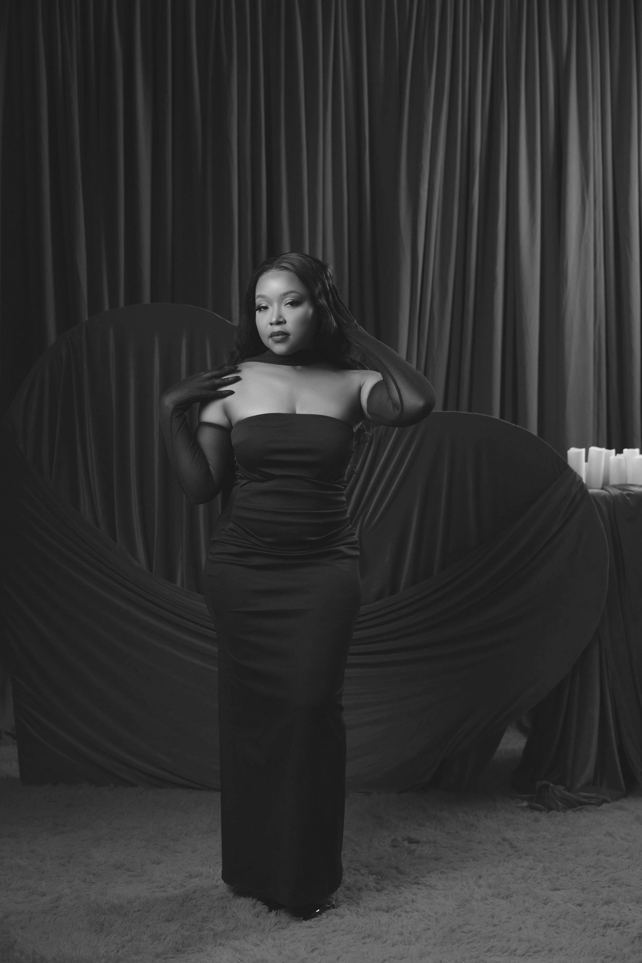 A woman in a strapless dress and gloves poses in front of fabric drapes, standing on a carpeted floor.