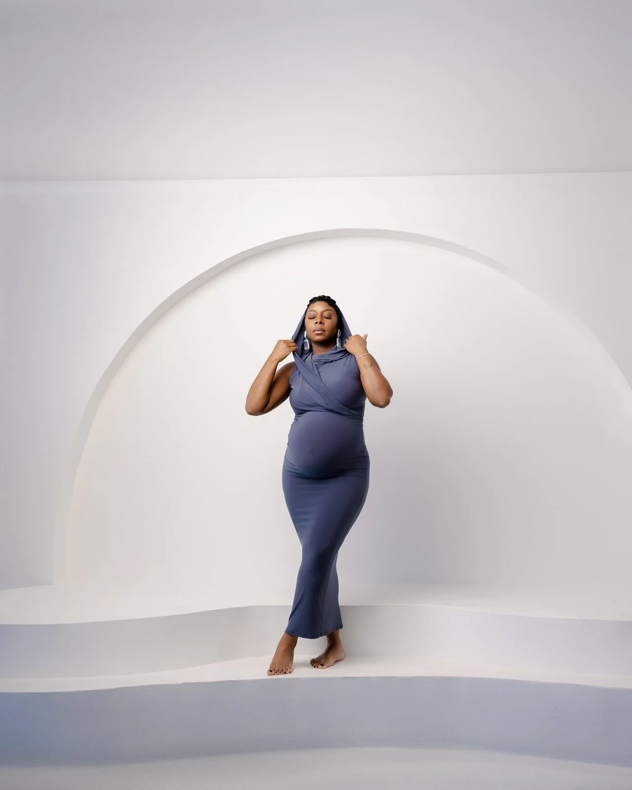 A woman in a form-fitting, sleeveless, dark blue dress standing barefoot in a minimalist, white studio with an arched wall design, eyes closed, adjusting her hooded dress.